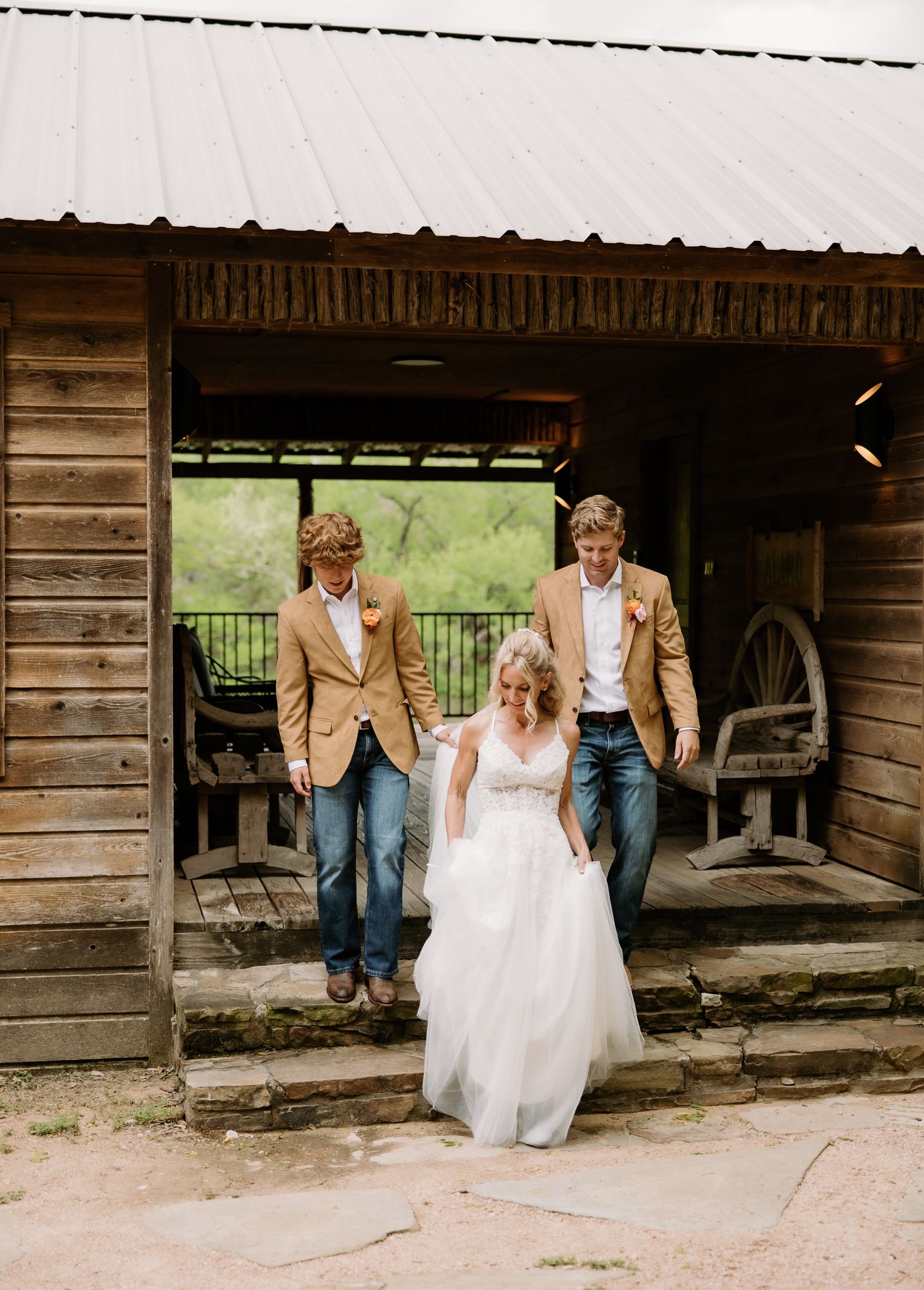 Bride in white gown walks with two groomsmen in tan blazers and jeans from a rustic wooden barn venue.