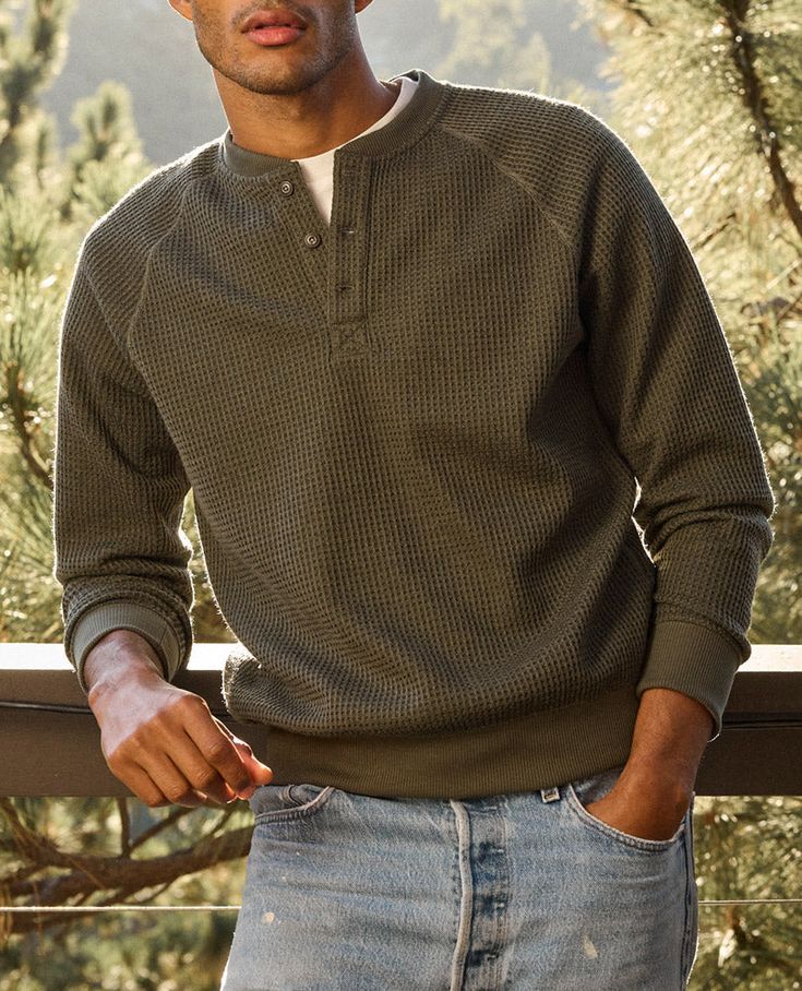Man wearing olive green waffle-knit henley sweatshirt with jeans standing on an outdoor wooden deck.