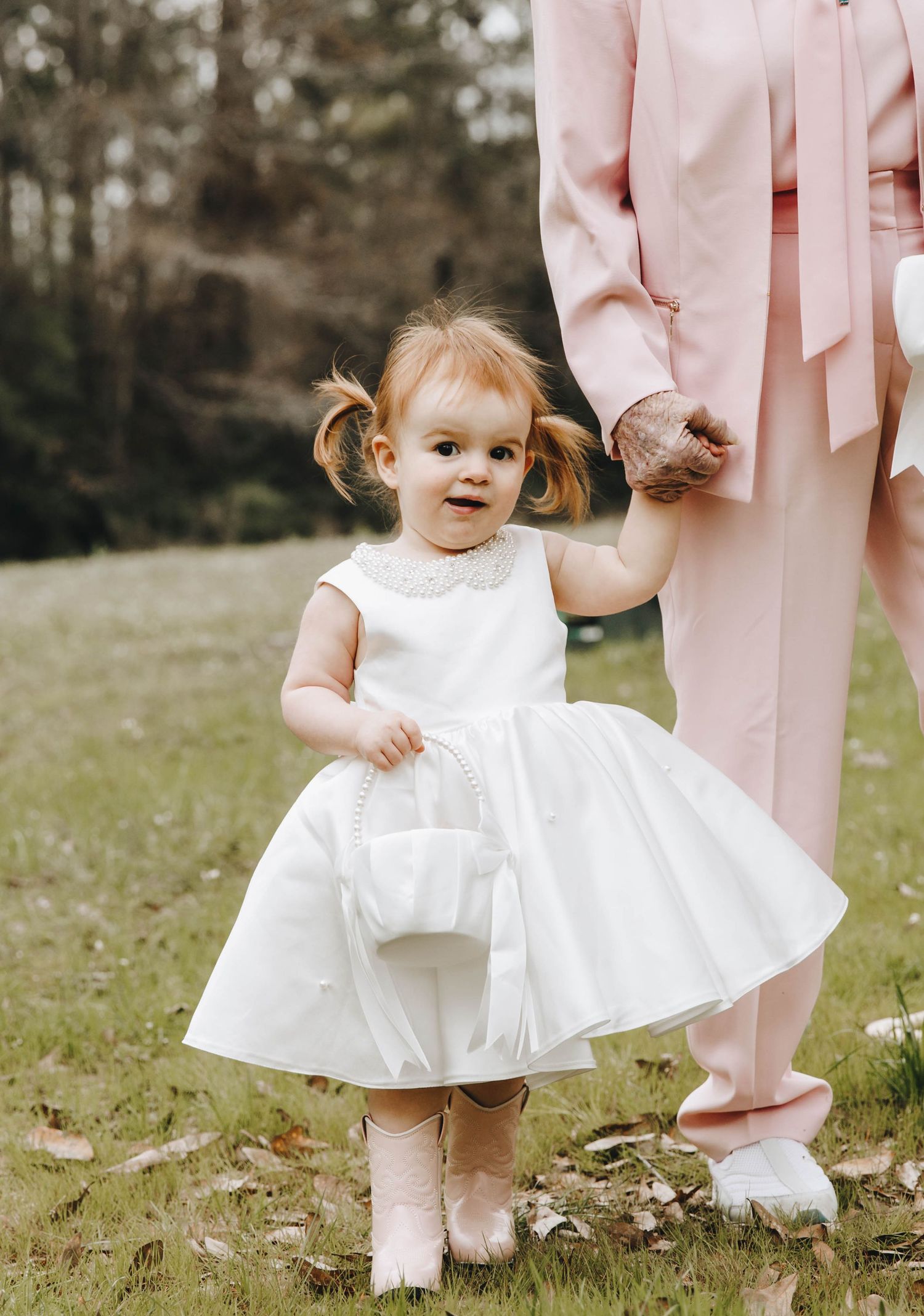 Adorable toddler flower girl in white dress and pink boots holds basket while standing on grass.