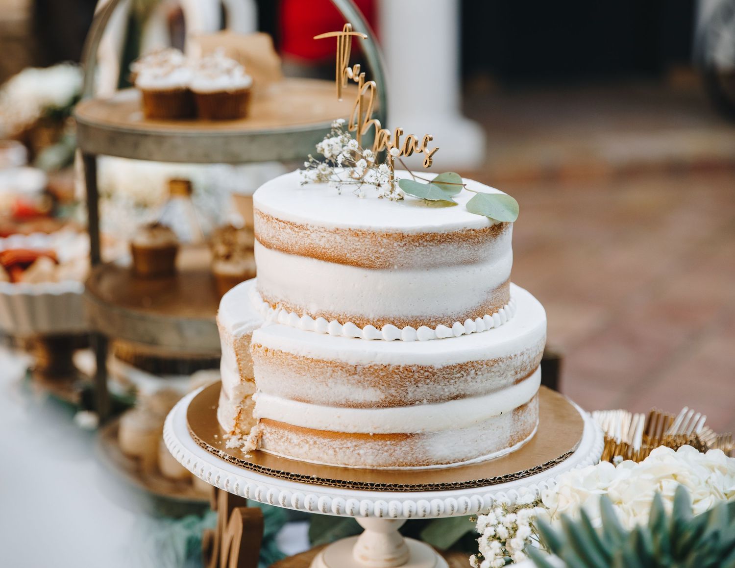 Two-tier naked wedding cake with white frosting, baby's breath, and a gold name topper on a dessert table.