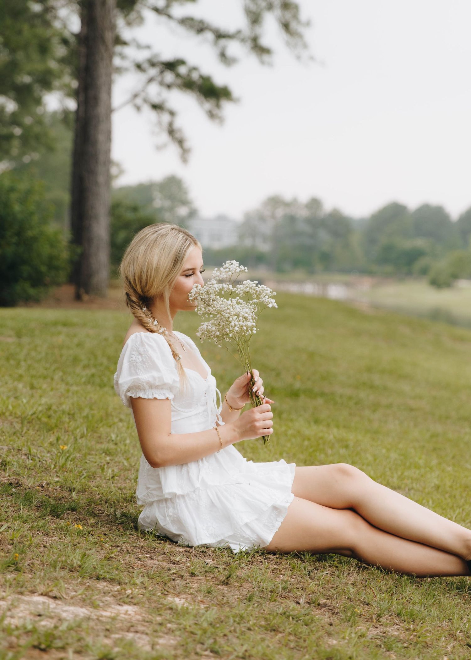 Blonde woman in white dress sitting on grass smelling a bouquet of wildflowers near a tree.
