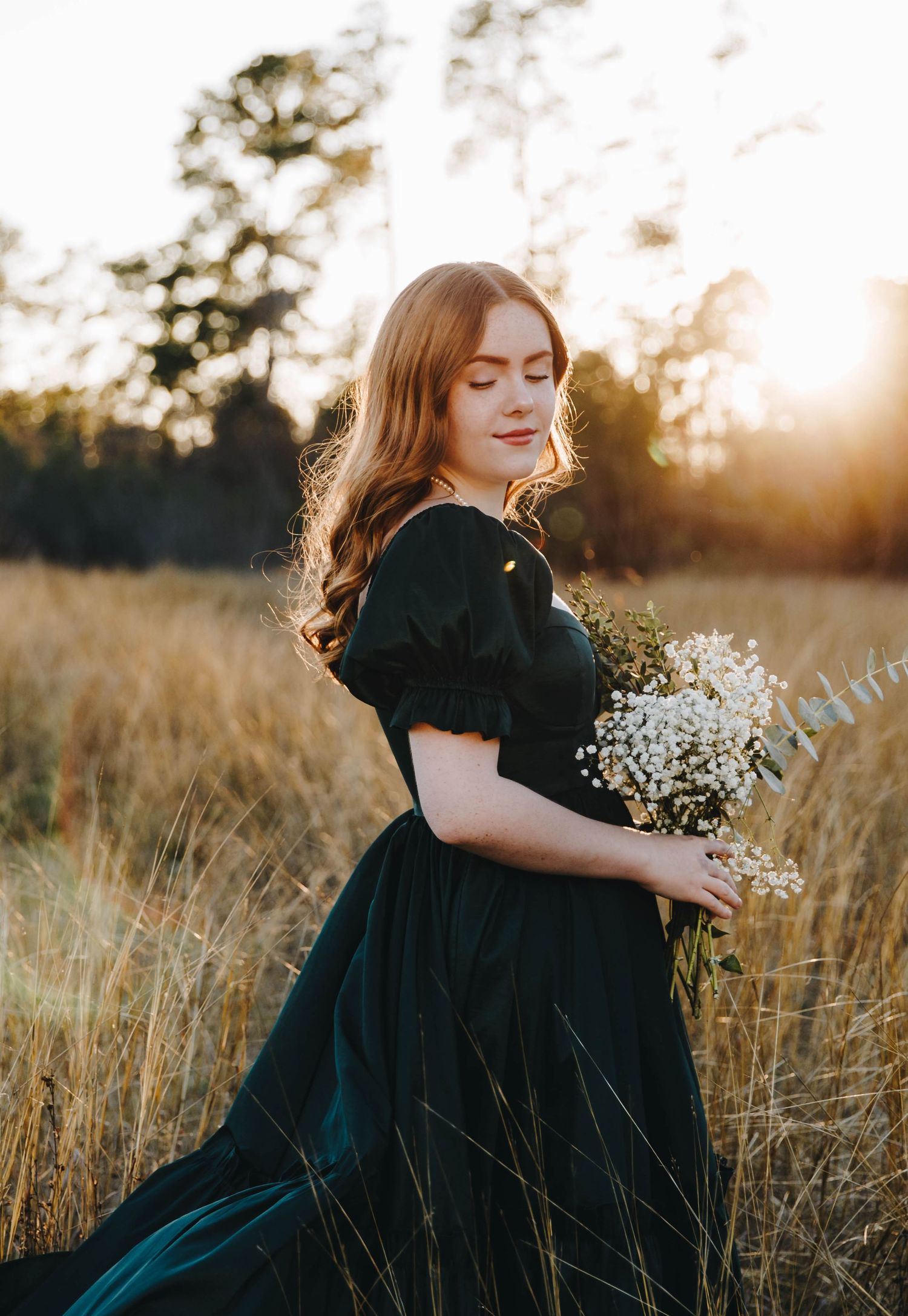 Young woman in dark green dress holding white flowers in a sunlit golden field at dusk.