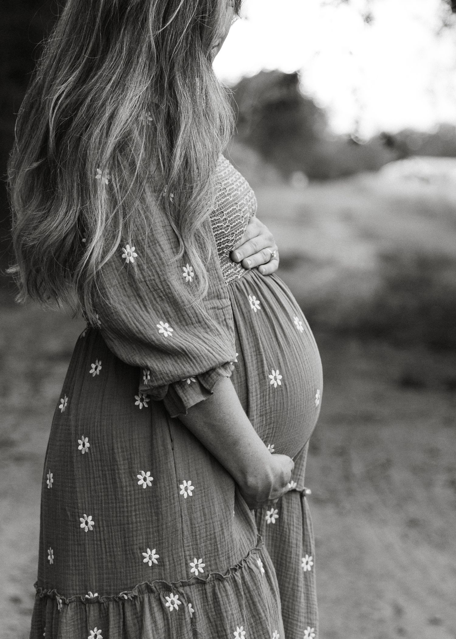 Black and white profile shot of pregnant woman in floral dress cradling baby bump outdoors.