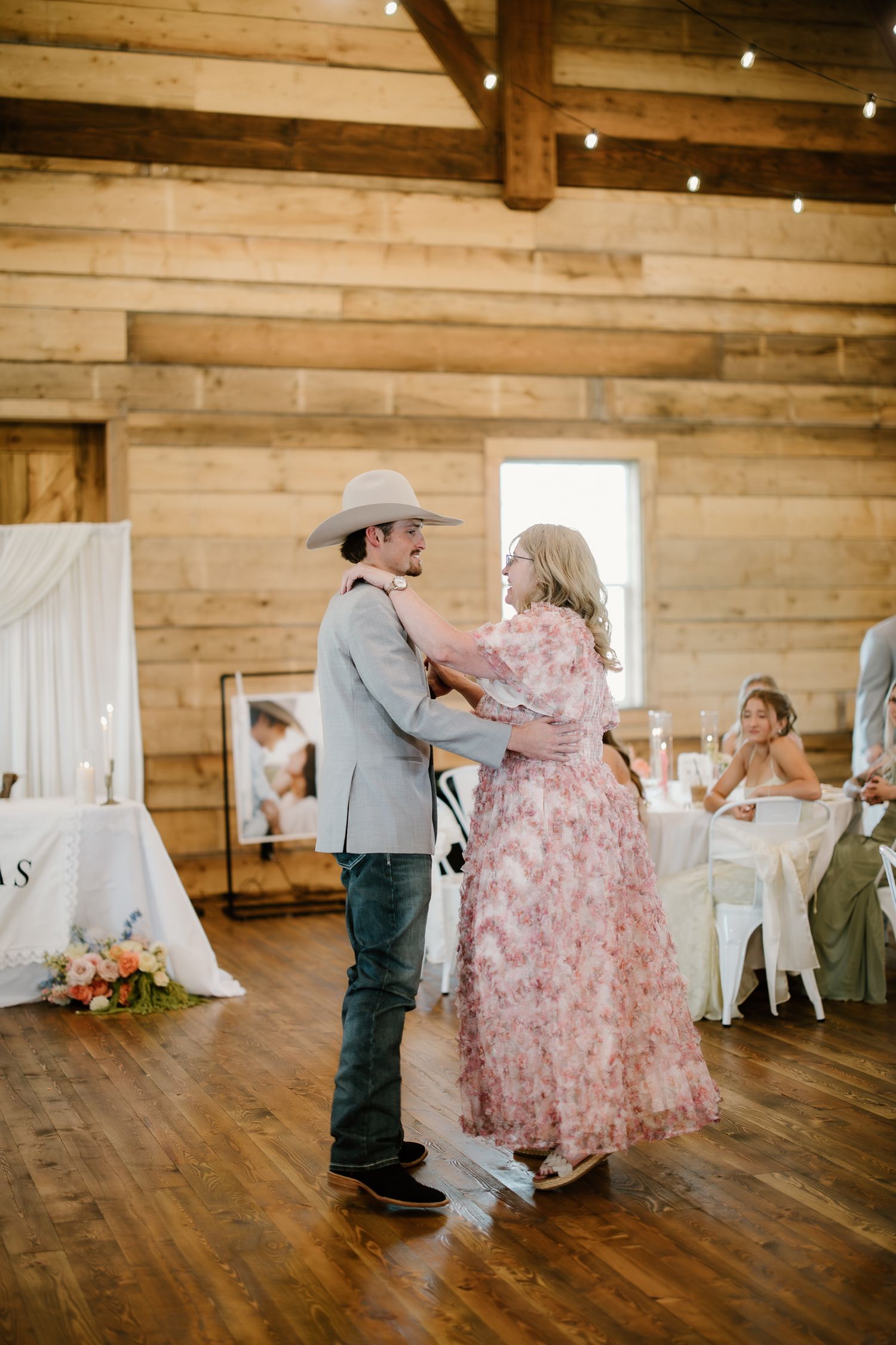 Groom in cowboy hat dancing with mother in floral dress inside rustic wooden barn reception venue.