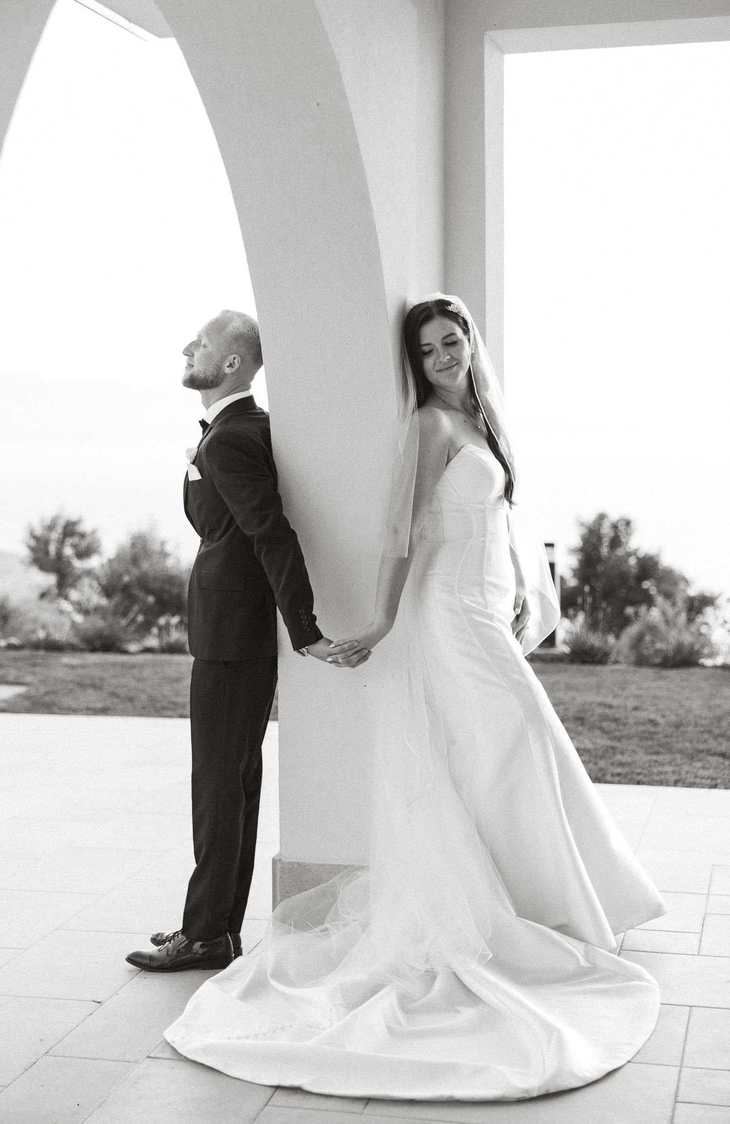 Bride and groom hold hands around a white column in a romantic black and white wedding portrait.