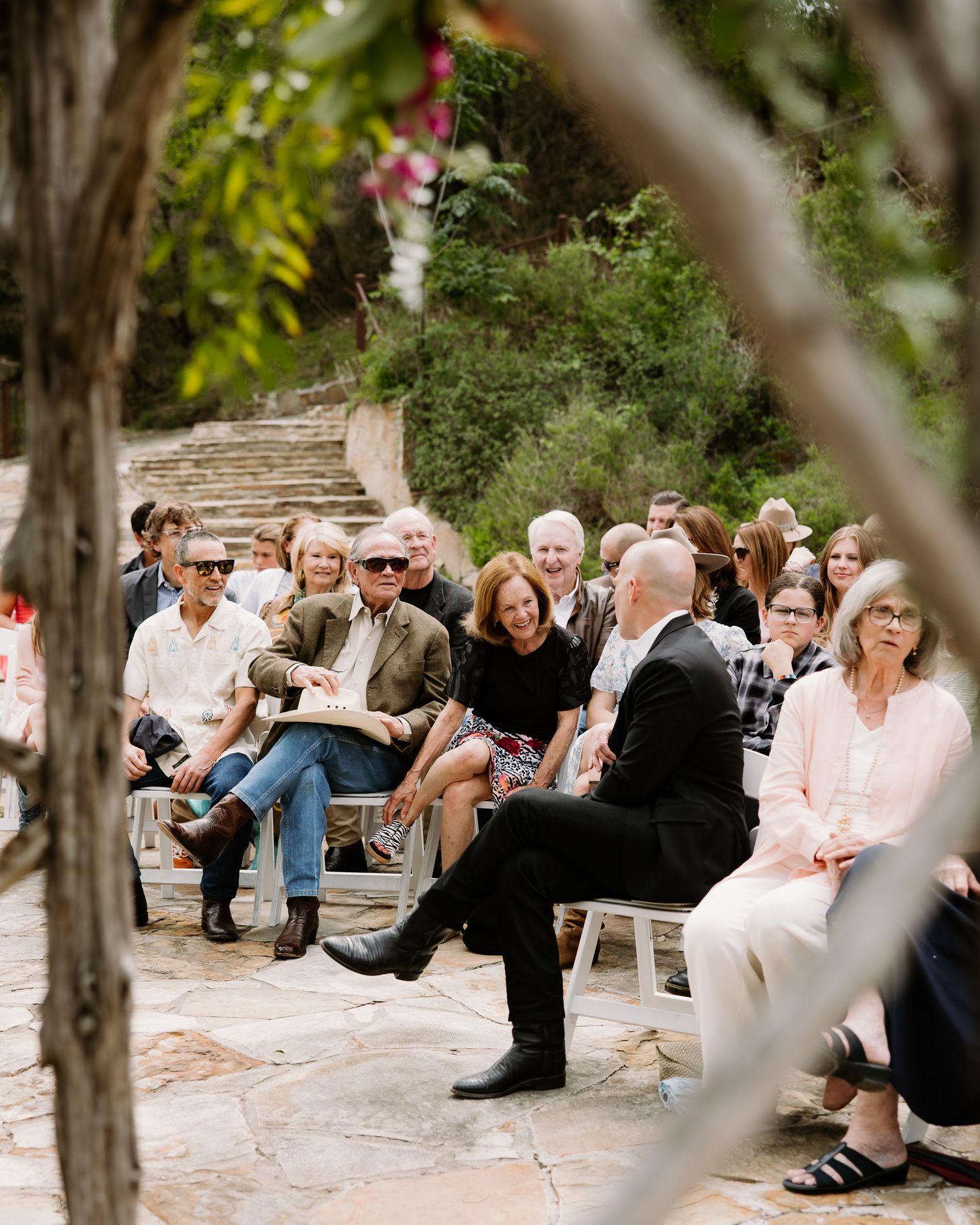 Wedding guests seated on white benches in an outdoor garden ceremony surrounded by lush greenery and stone pavers.