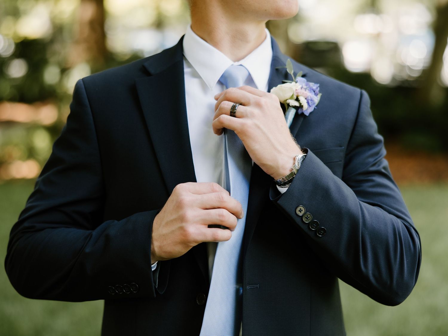 Groom in navy suit adjusting light blue tie with boutonniere, preparing for wedding ceremony outdoors.
