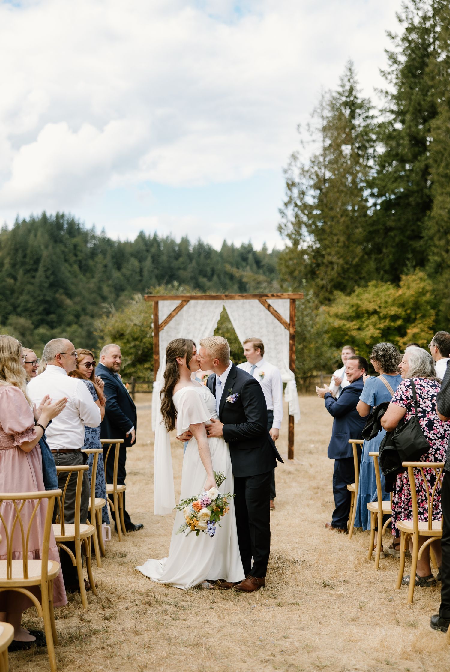 Bride and groom share first kiss as married couple while guests cheer in outdoor ceremony aisle.