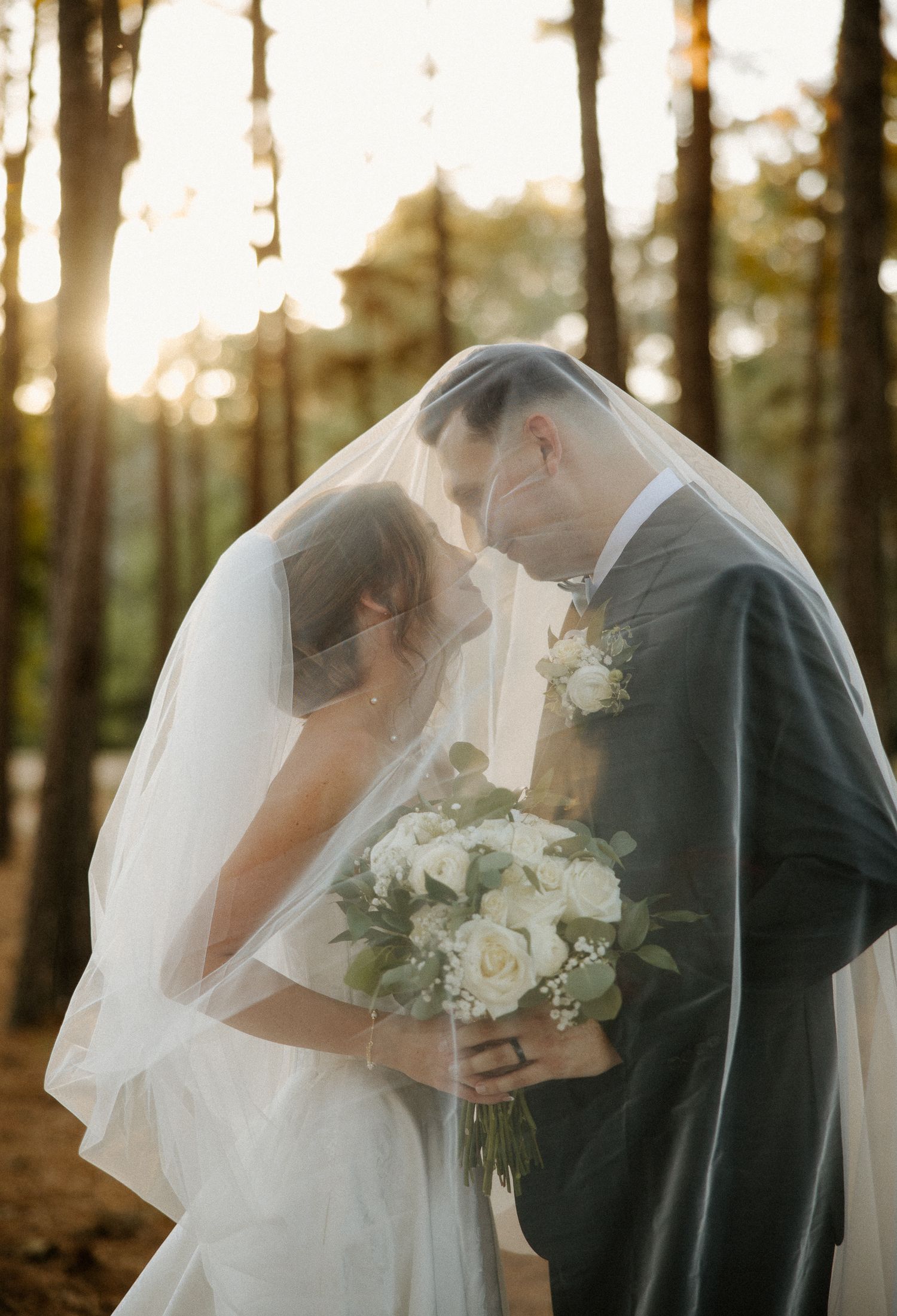 Bride and groom sharing intimate moment under wedding veil in sunlit forest setting.