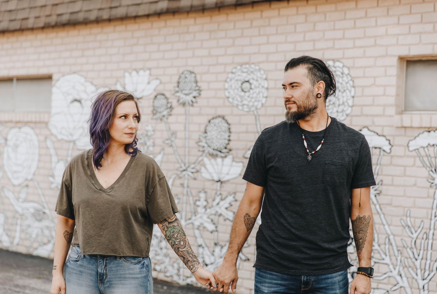 Tattooed couple holding hands while walking past a floral mural on a brick wall.