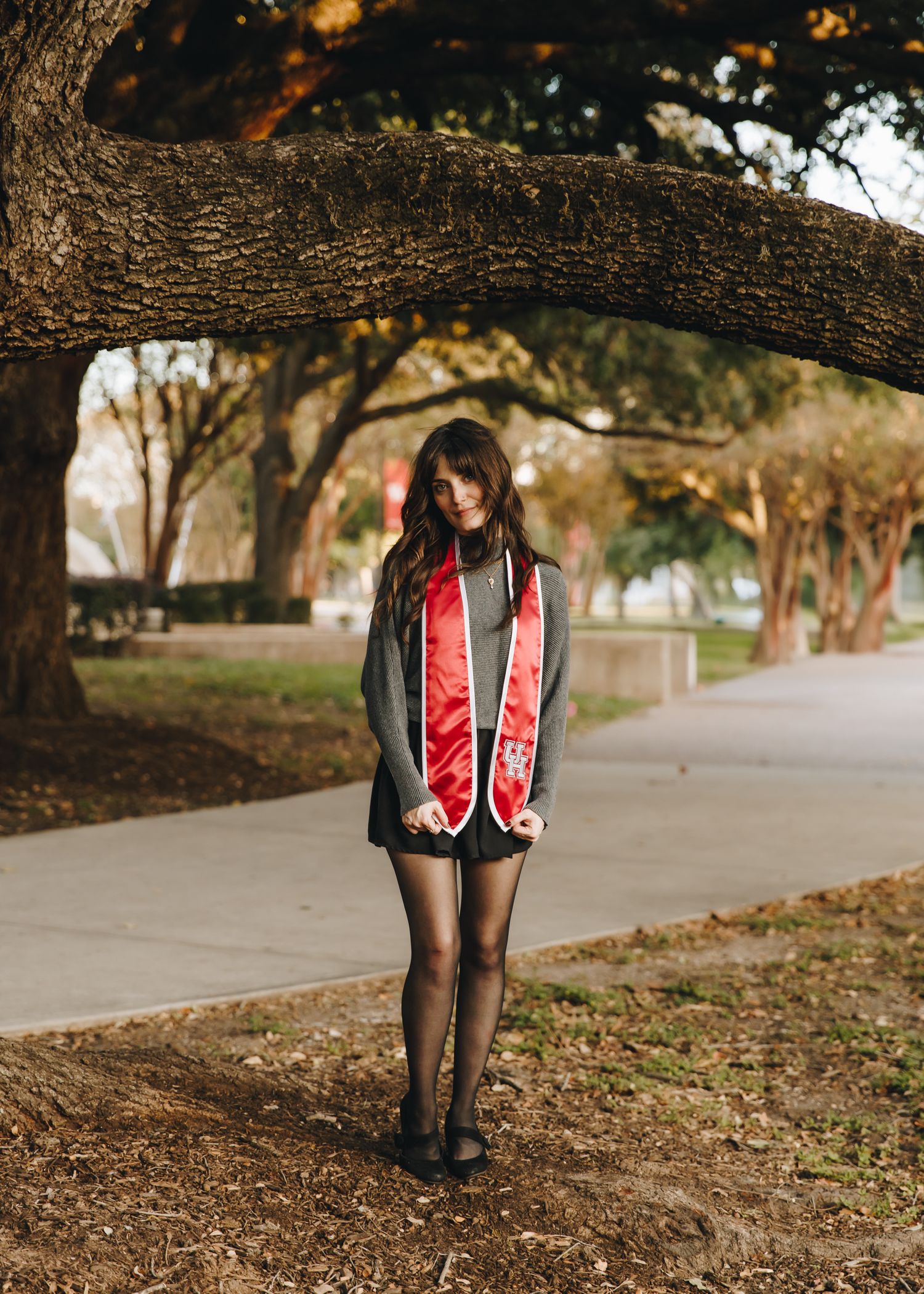 Young woman in mini dress wearing red graduation stole standing under large oak tree on campus.