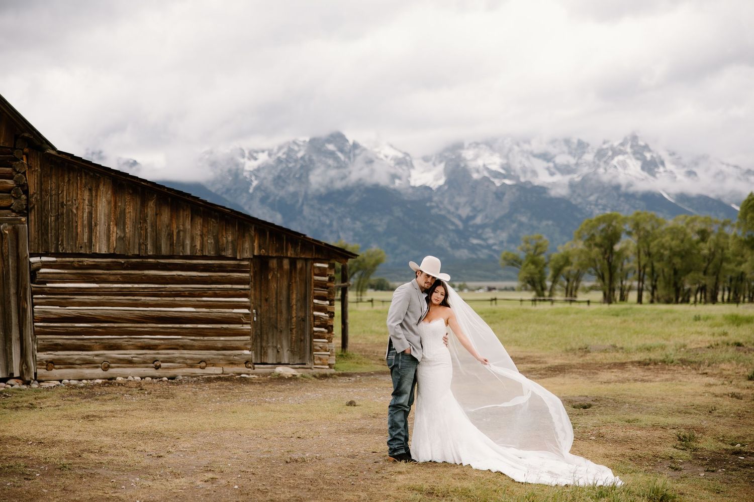 Bride and groom share a romantic kiss near a rustic barn with the Grand Teton mountains in the background.