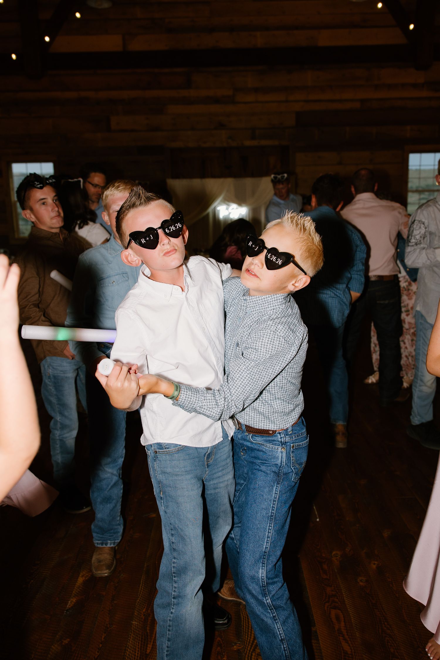 Two young boys wearing heart-shaped sunglasses dancing with glow sticks at wedding reception.