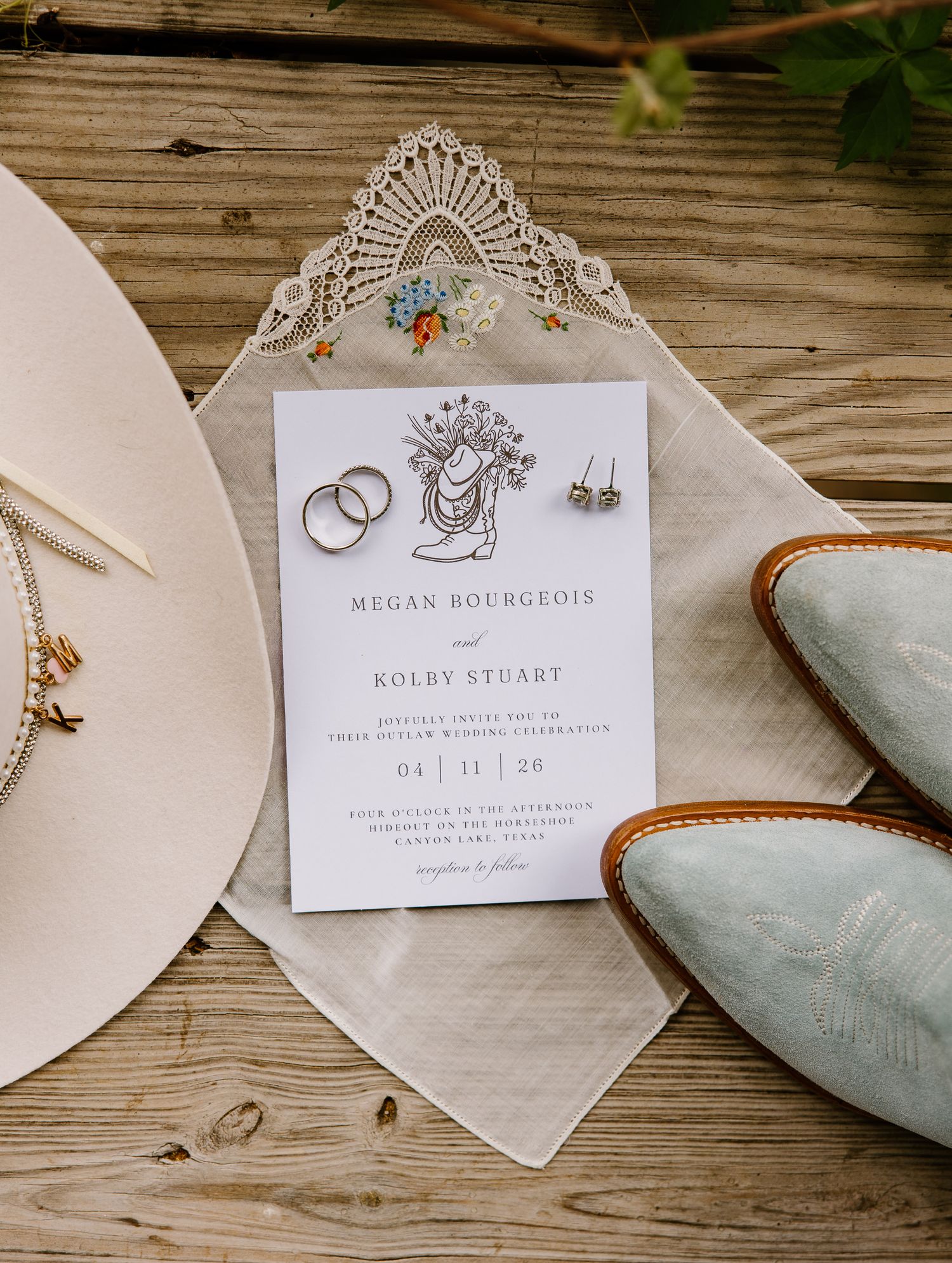 Rustic western wedding invitation for Megan Bourgeois and Kolby Stuart with cowboy boots, gold rings, and earrings on wood.