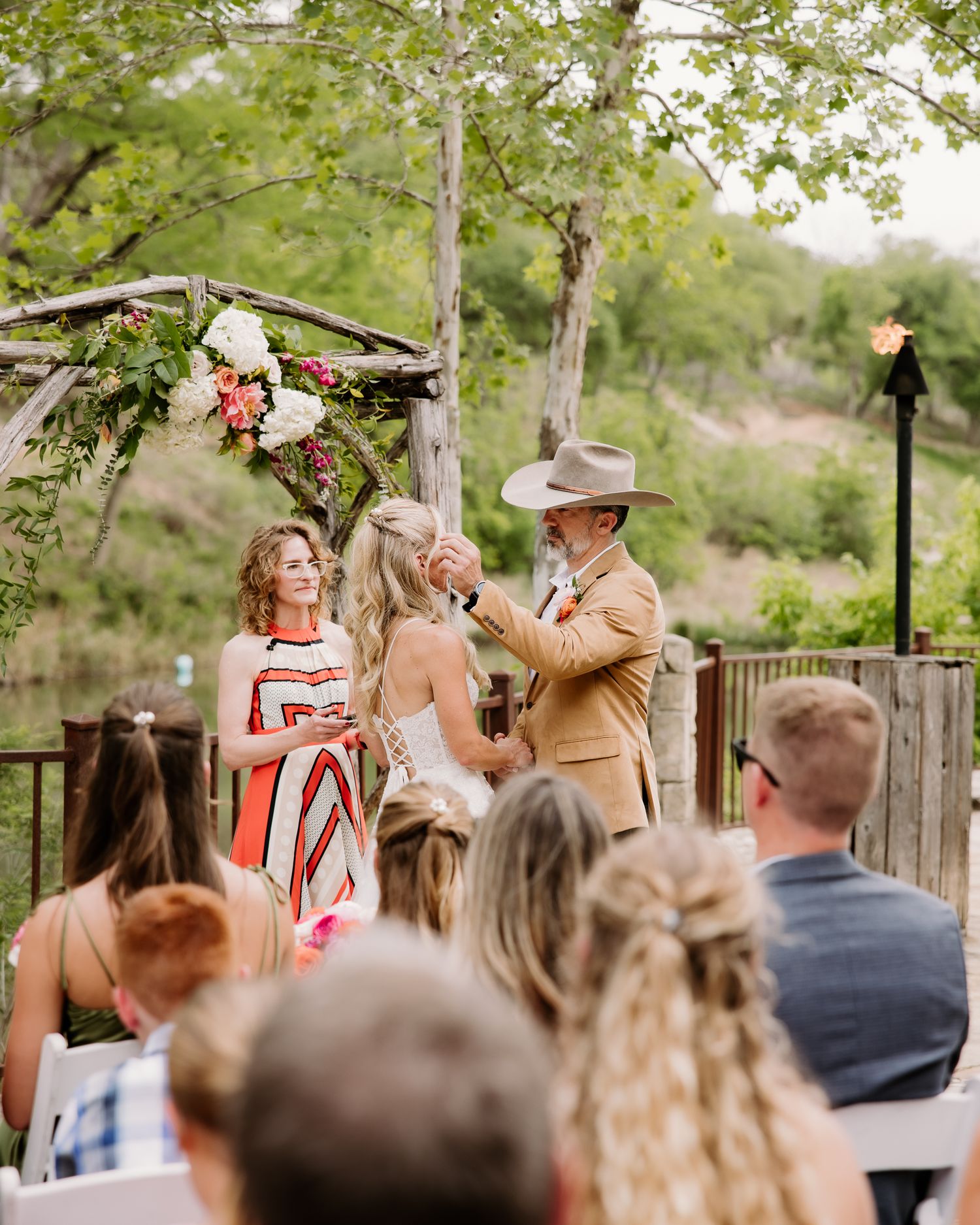 Outdoor wedding ceremony under floral arch with officiant and groom in tan suit lifting bride's veil.