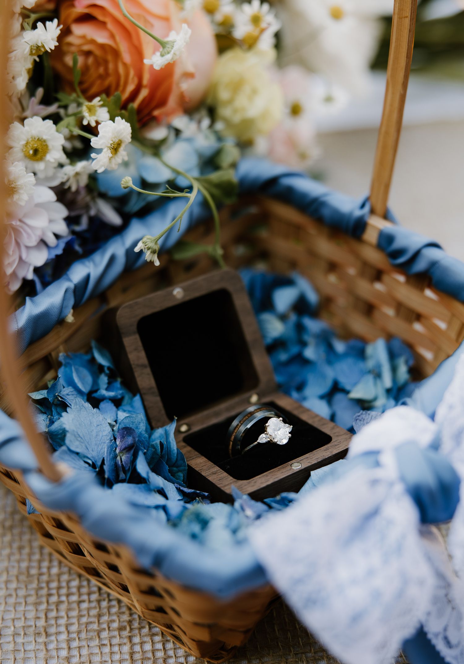 Wedding rings displayed in a wooden box with blue hydrangeas and floral bouquet in a wicker basket.