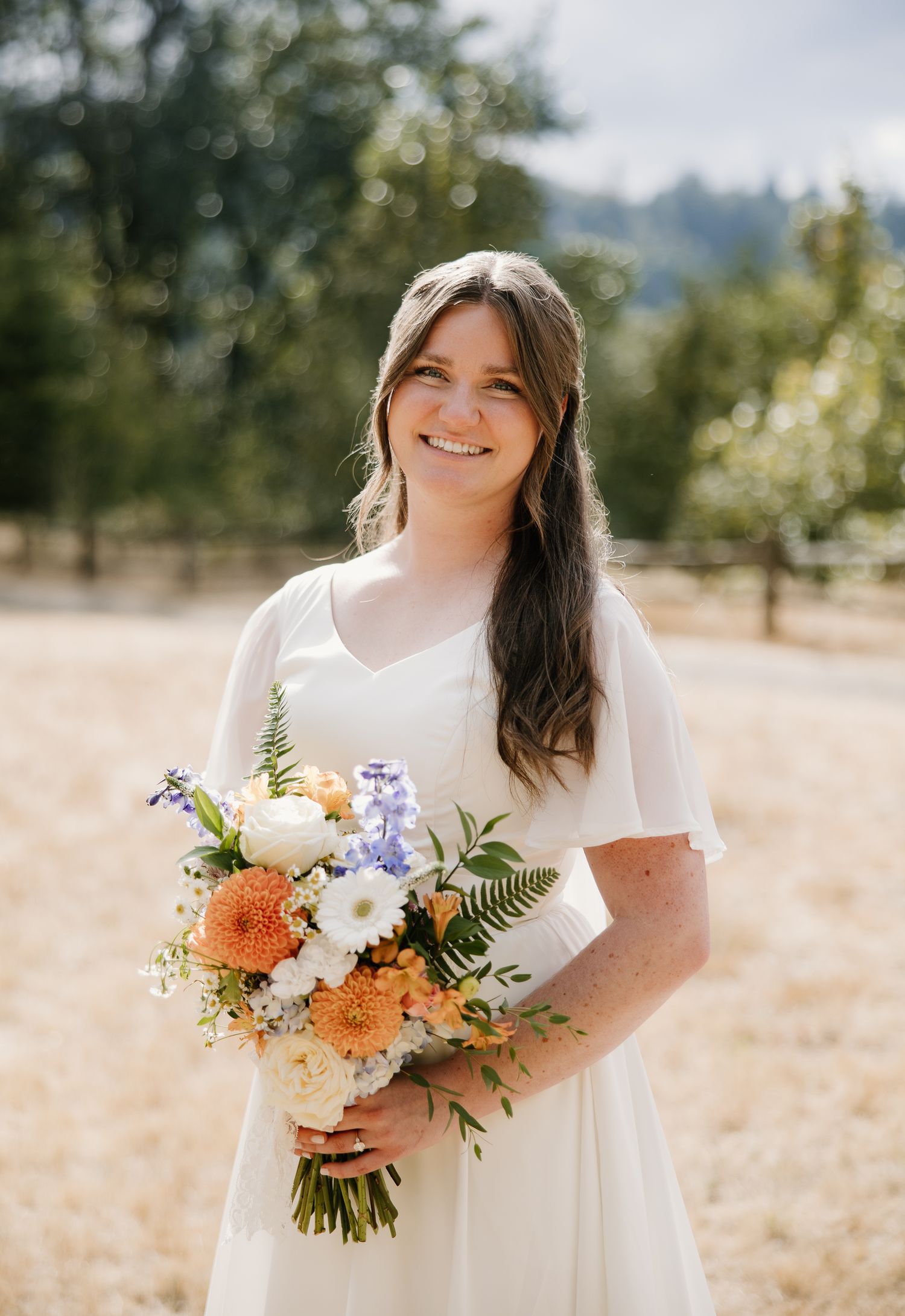 Smiling bride in white dress holds colorful orange and white bouquet outdoors.