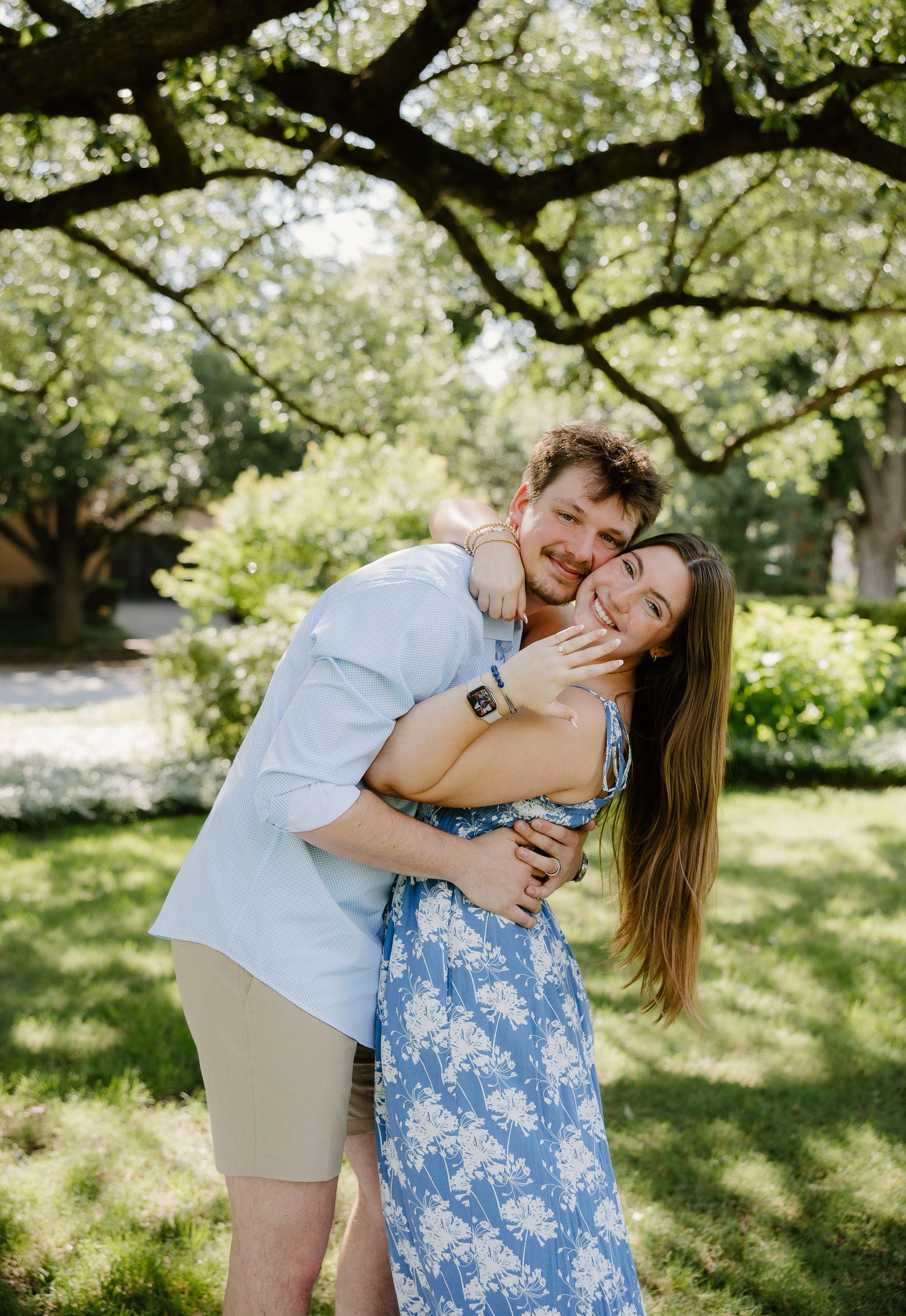 Man hugging woman from behind under oak trees, both smiling with engagement ring visible.