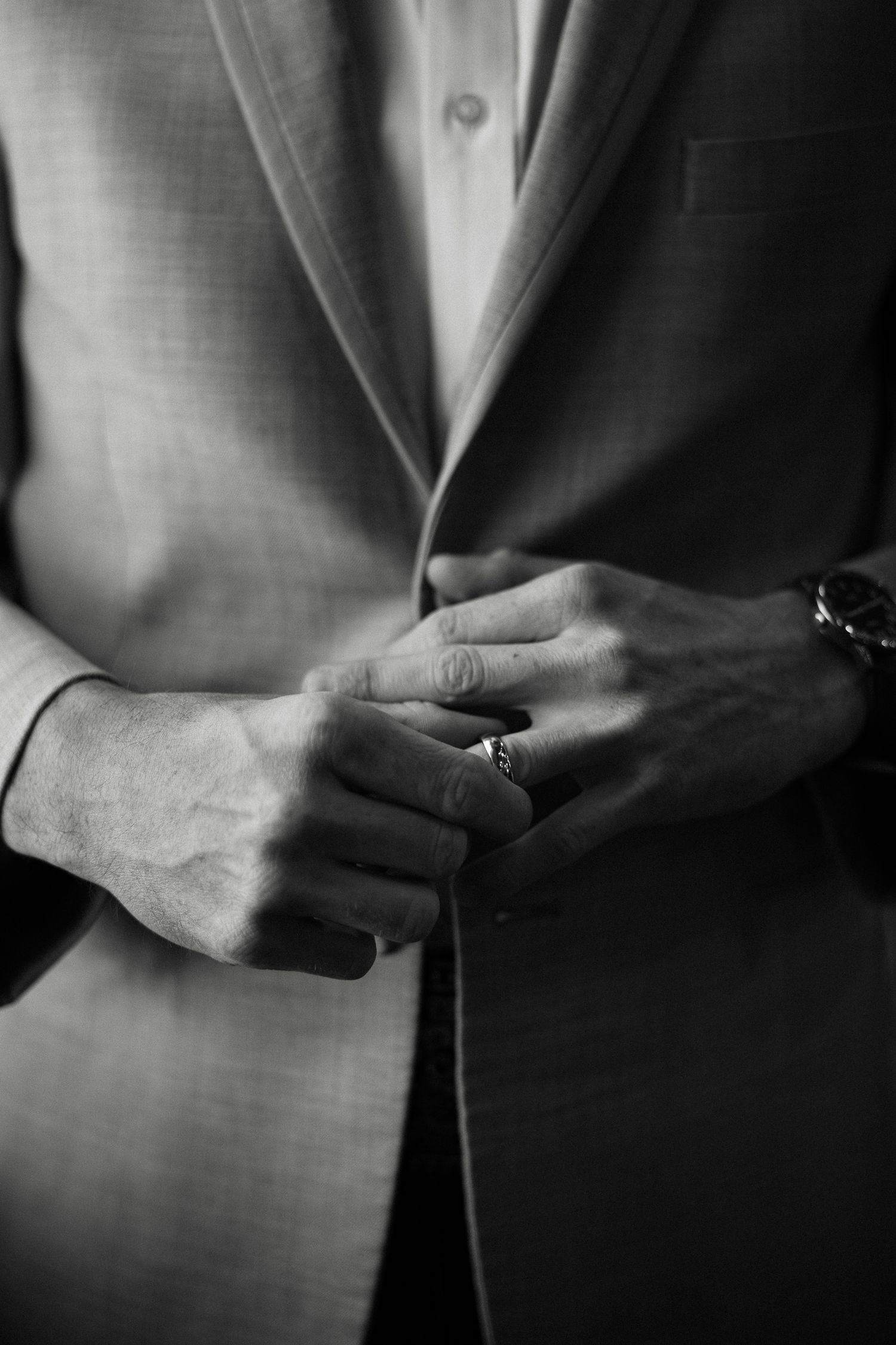 Black and white close-up of groom adjusting suit jacket with wedding ring and watch visible.
