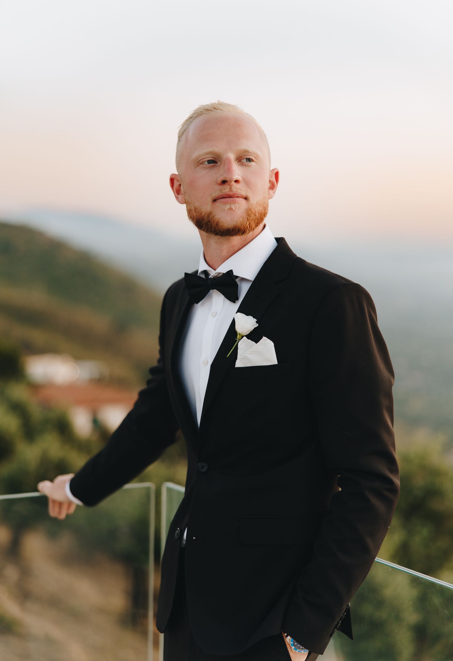 Groom in black tuxedo with bow tie and white boutonniere poses on balcony with scenic mountain views at sunset.