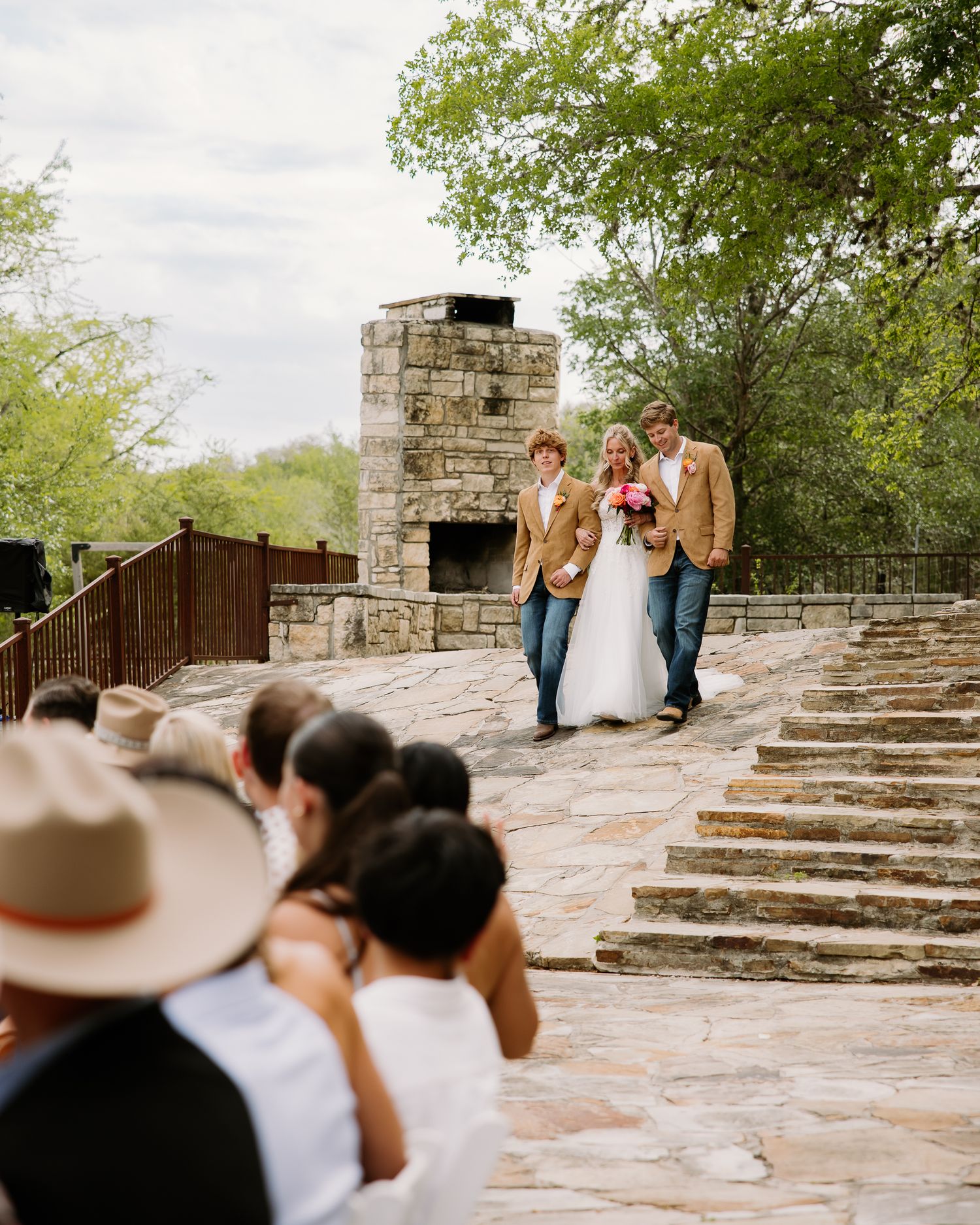 A bride in white is escorted down stone steps by two people in matching tan blazers at an outdoor wedding ceremony.