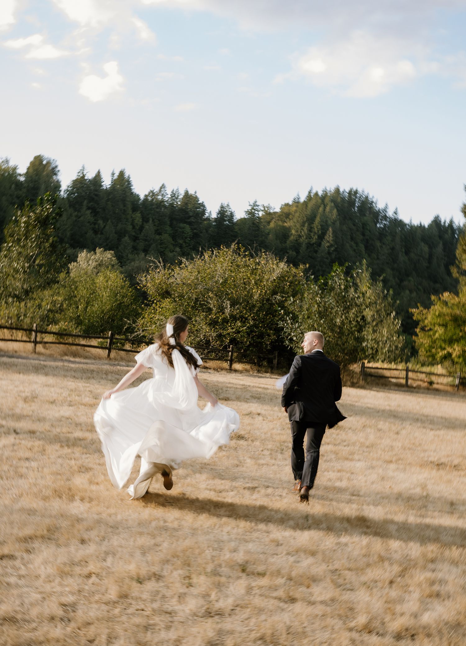 Bride in flowing white gown and groom running joyfully through golden meadow with forest backdrop.