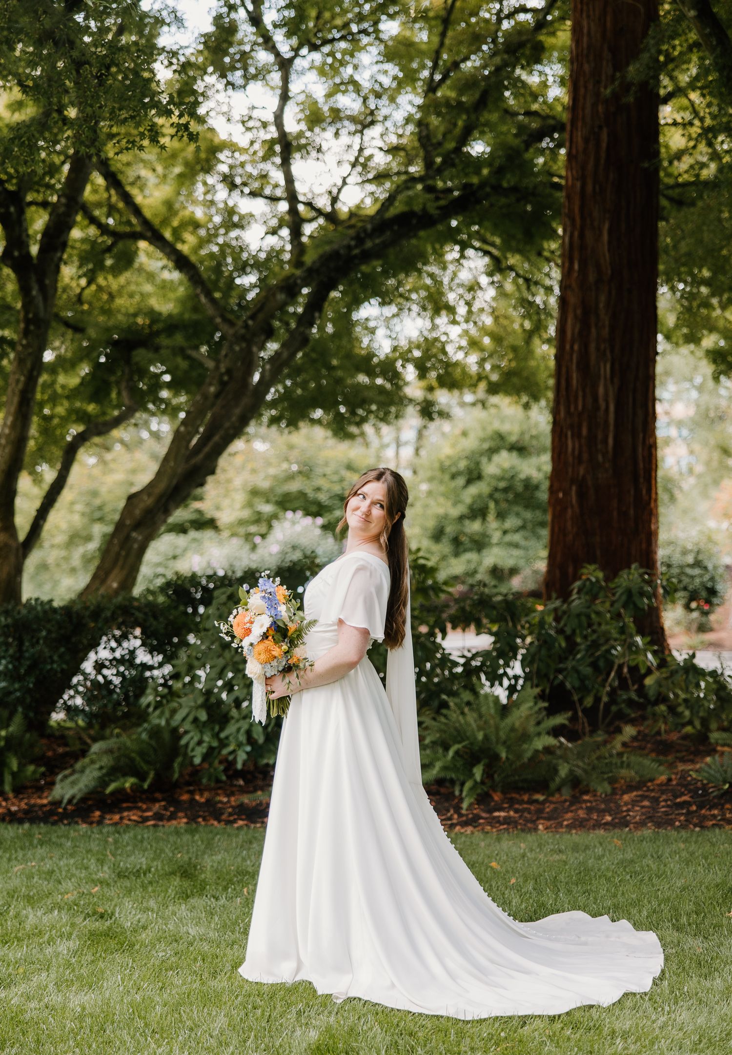 Bride in elegant white gown holds colorful bouquet while posing in a lush garden setting.