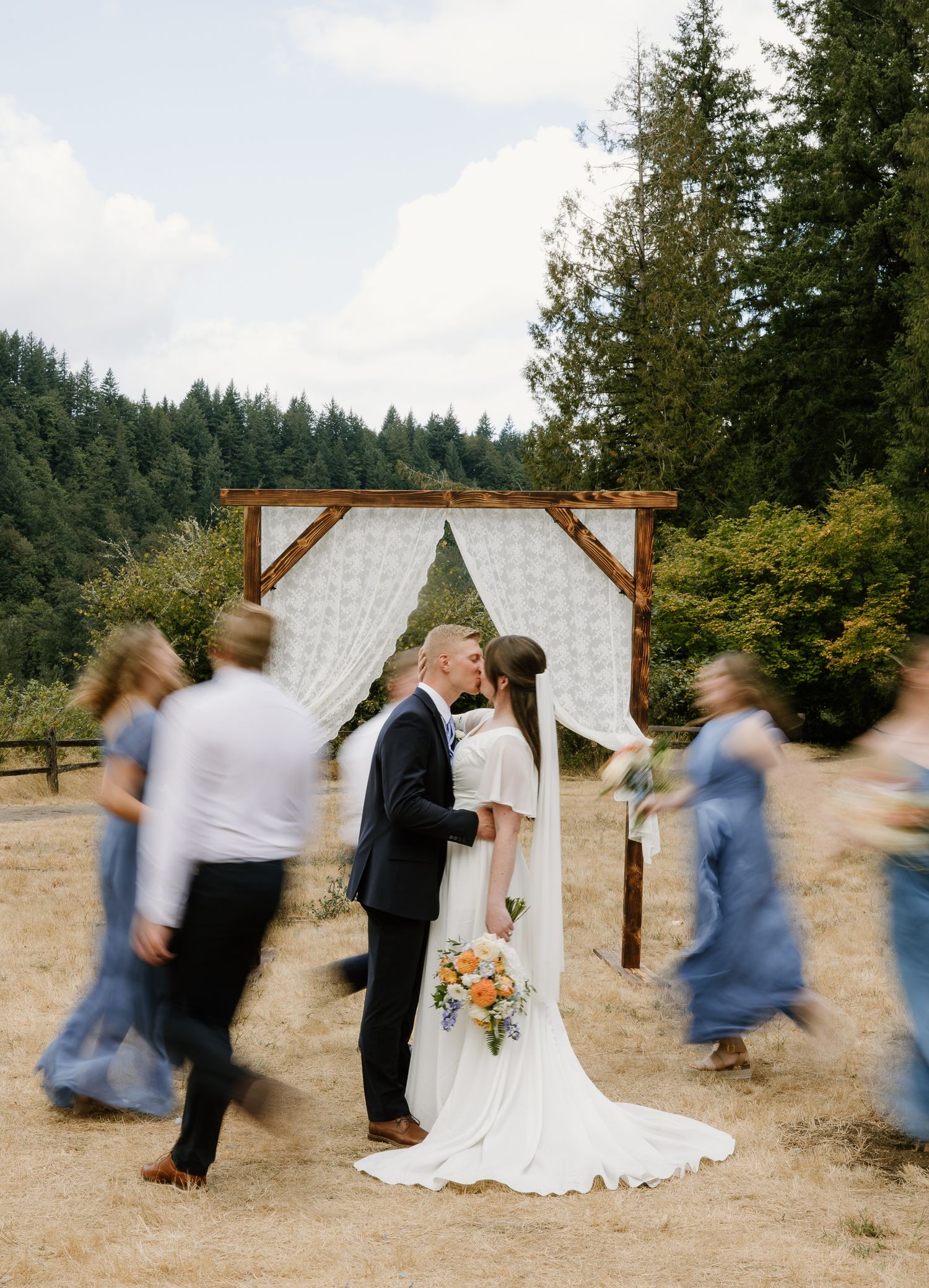 Bride and groom share first kiss under rustic wooden arch as wedding party walks past in blur.