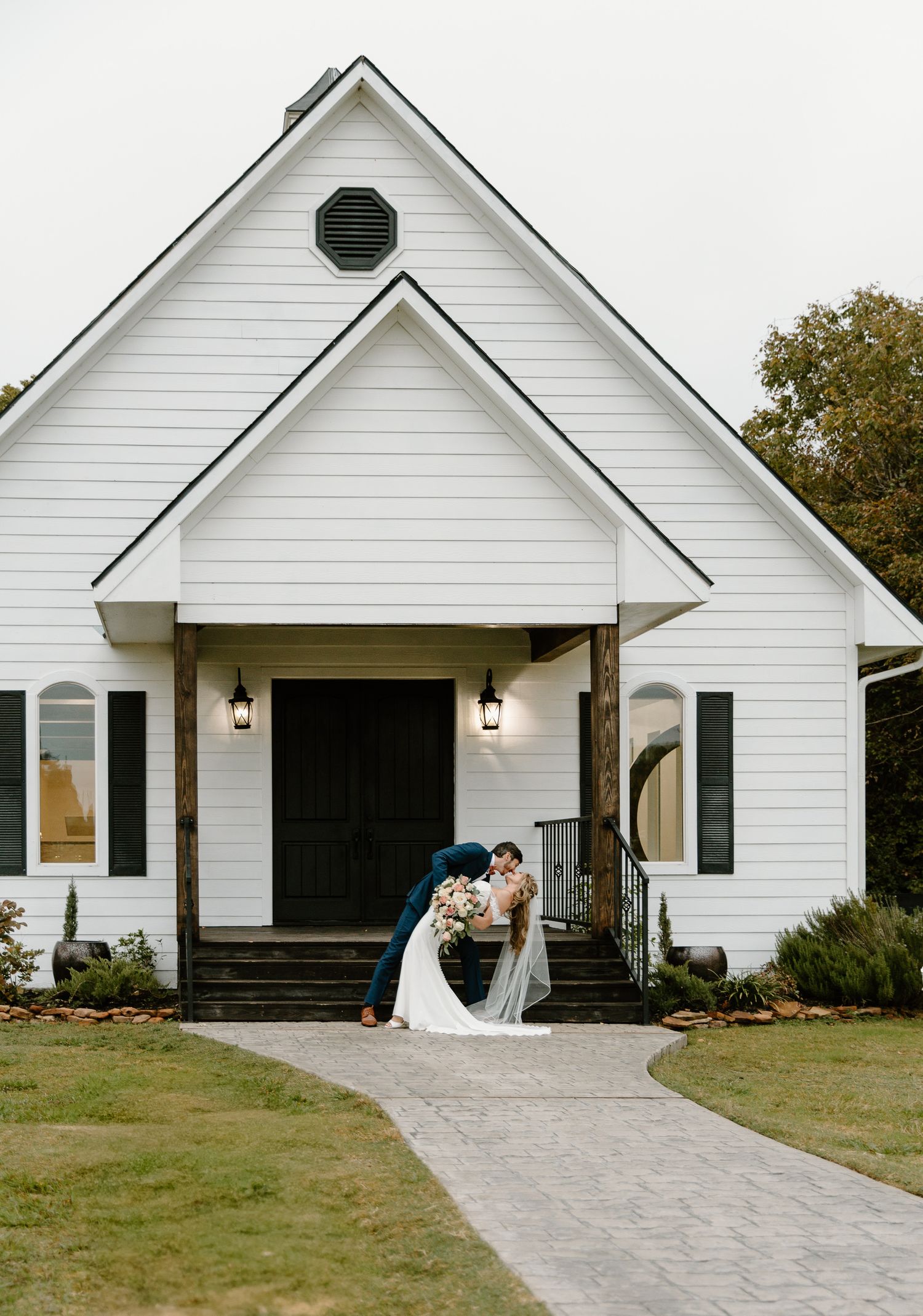 Bride and groom kissing in front of charming white chapel with dark shutters and stone pathway.