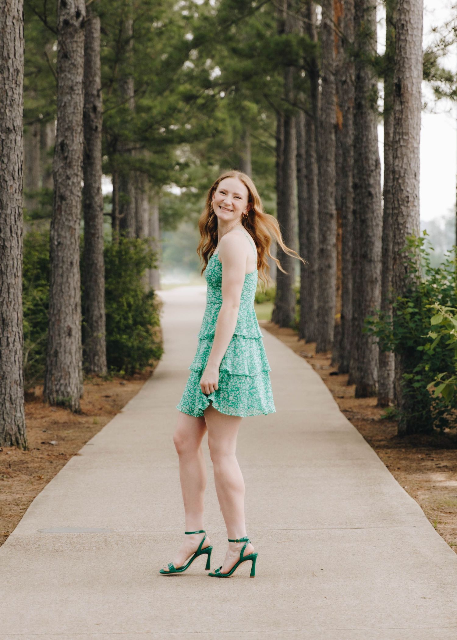 Young woman in green floral dress and heels posing on tree-lined pathway in elegant senior portrait photo.
