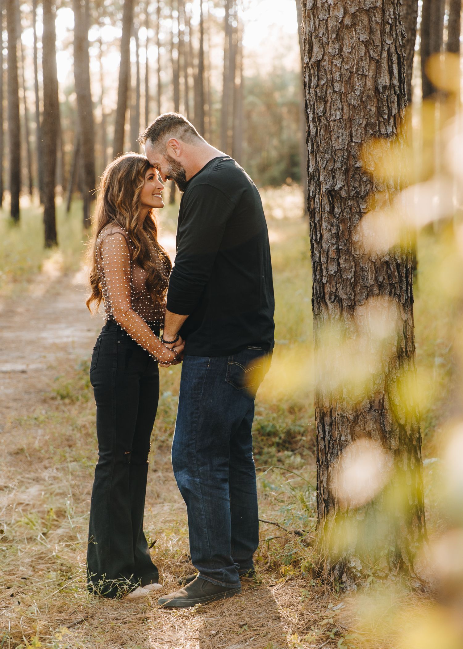 Couple holding hands and smiling on a wooded path with soft bokeh foliage in the foreground.