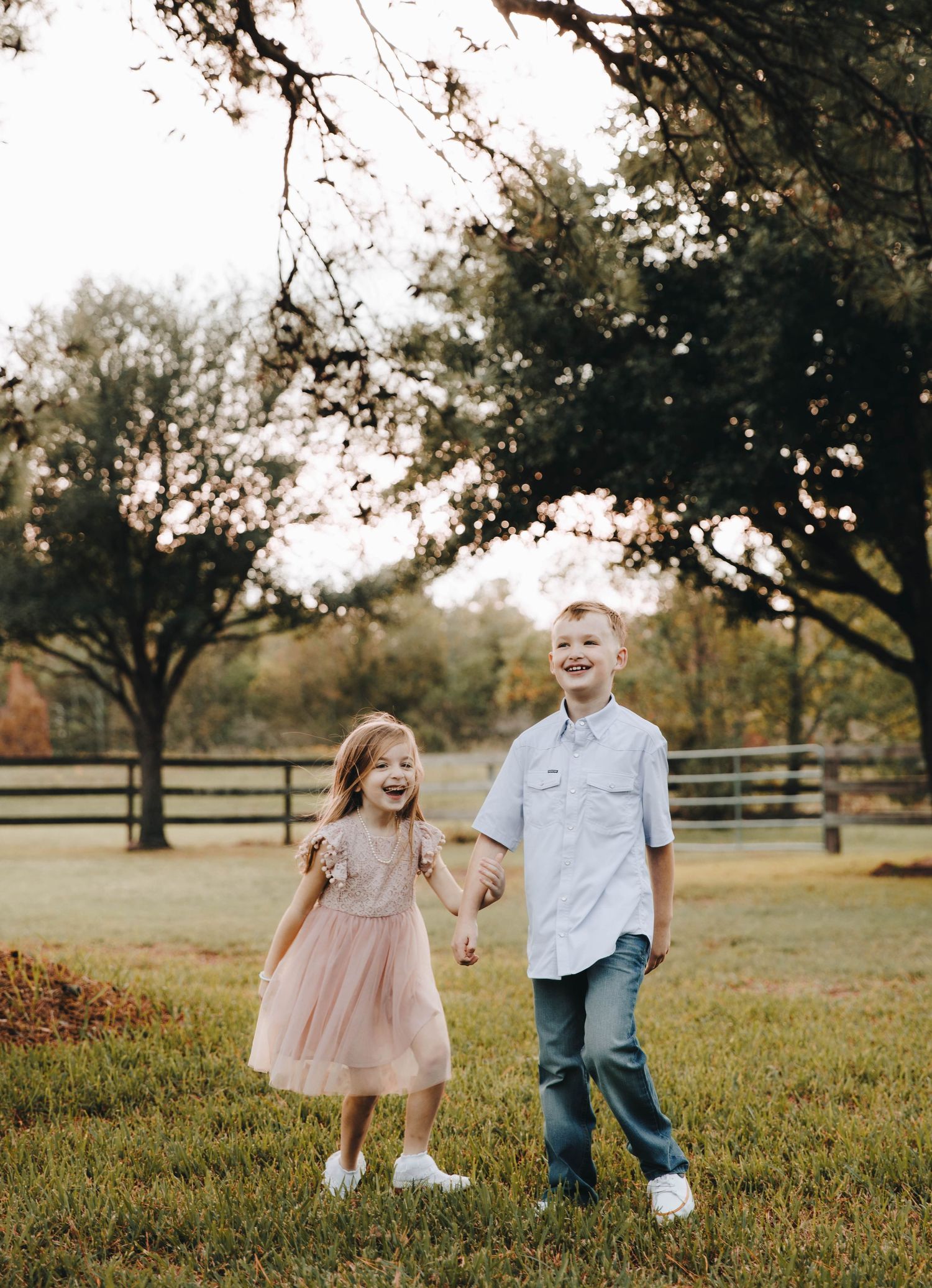 Two happy children holding hands and laughing in a green field during a golden hour outdoor photo session.