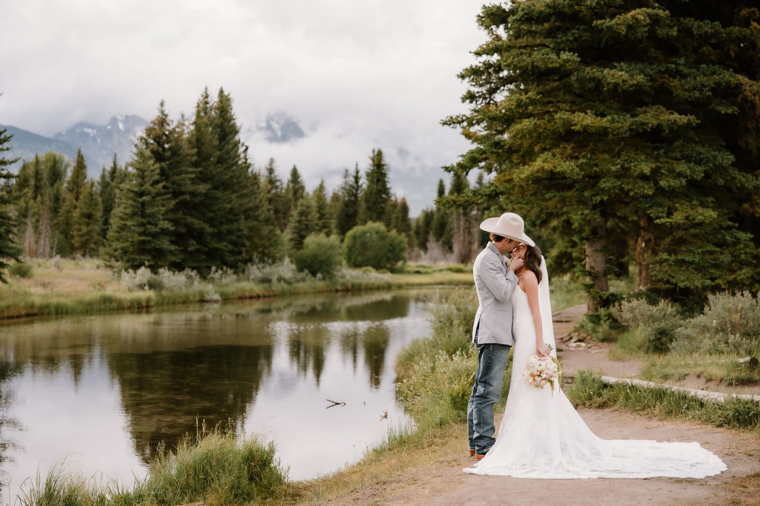 Bride and groom embracing by a mountain lake with evergreen trees and misty peaks in Grand Teton National Park.