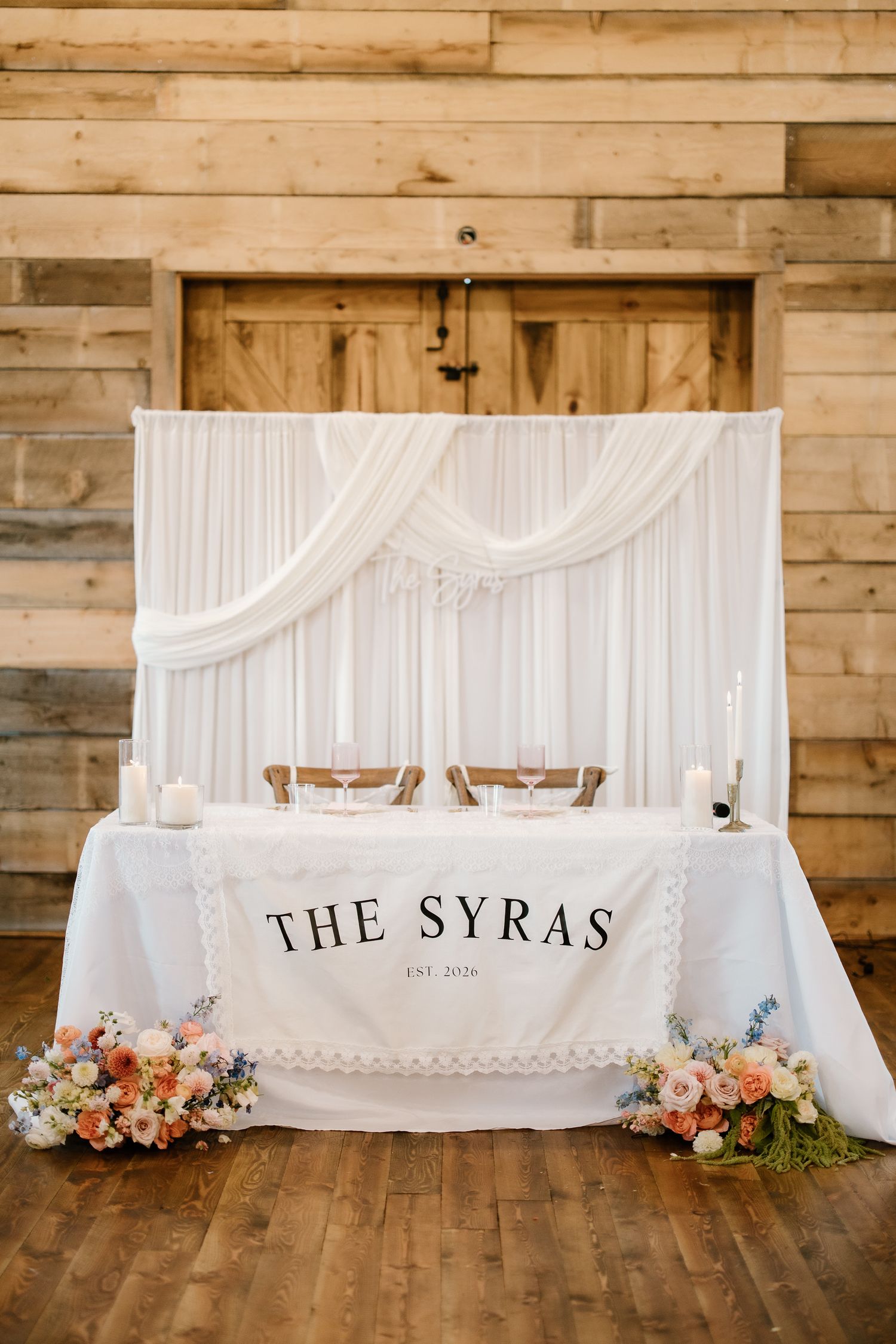 The Syras est. 2026 sweetheart table with white draping and floral arrangements in barn.