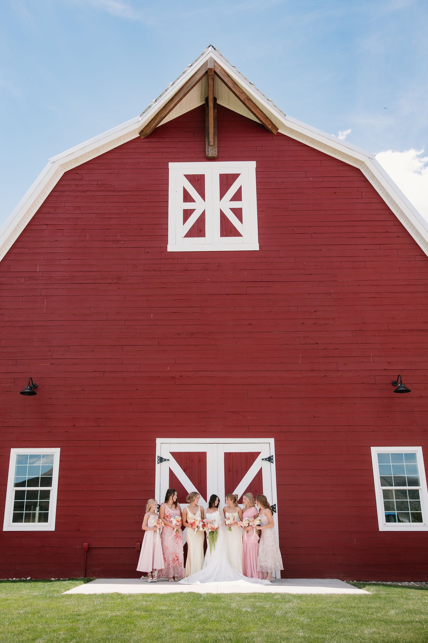 Bride and bridesmaids in pink dresses posing in front of a classic red barn on a sunny wedding day.