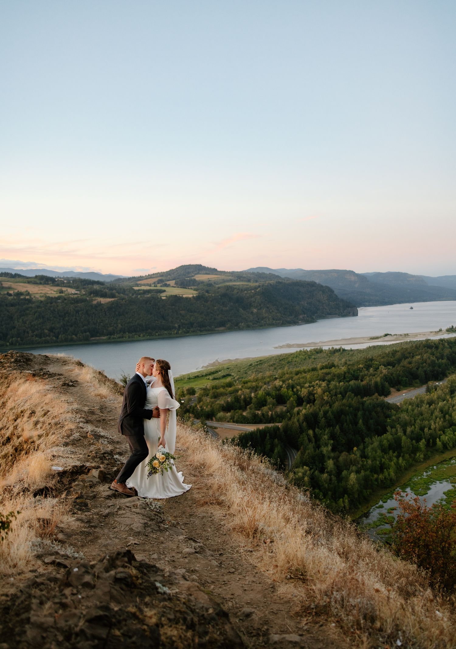A bride and groom share a kiss on a scenic hilltop overlooking the Columbia River Gorge at sunset.
