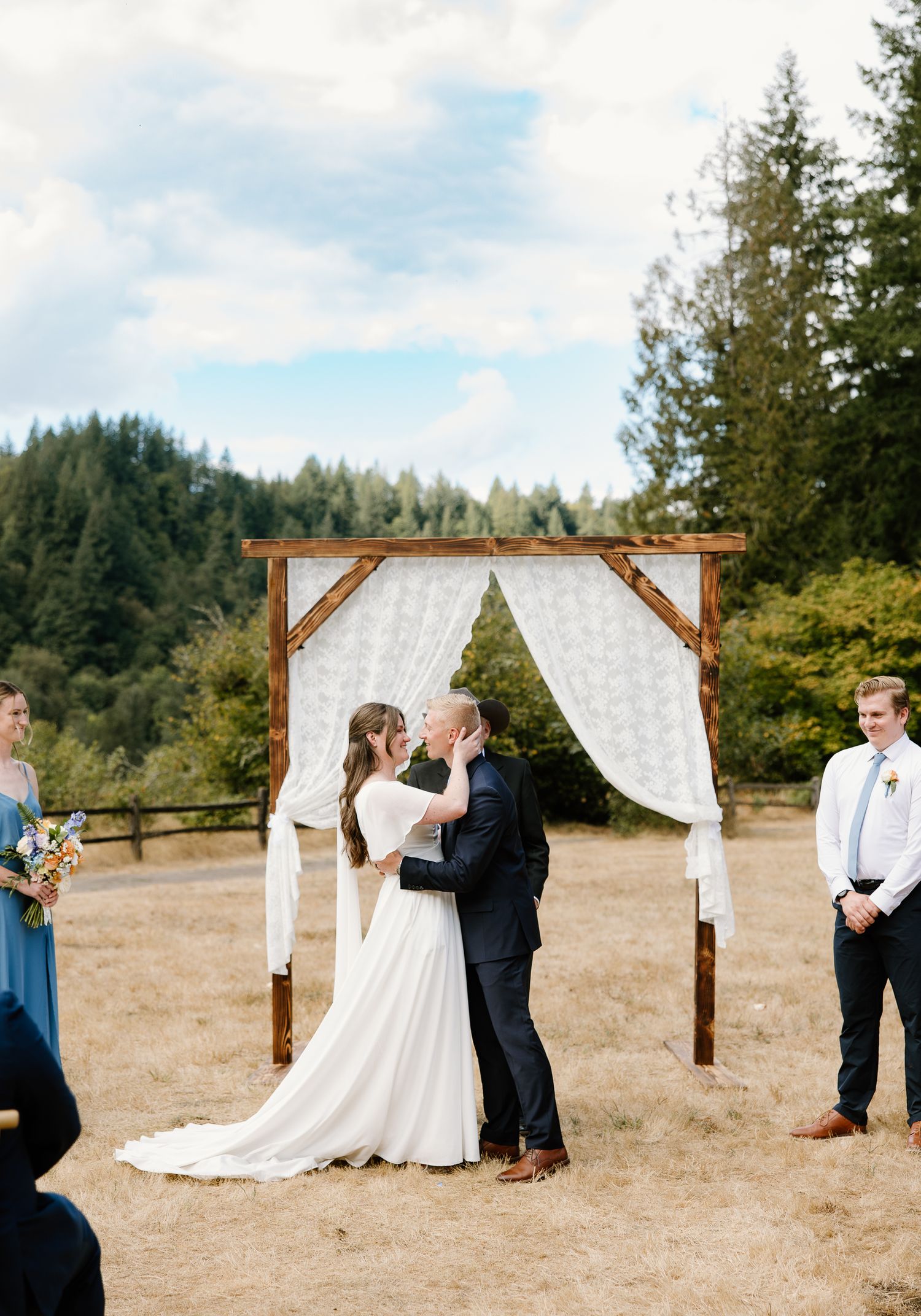 Bride and groom share their first kiss under a rustic wooden arch at an outdoor wedding ceremony.