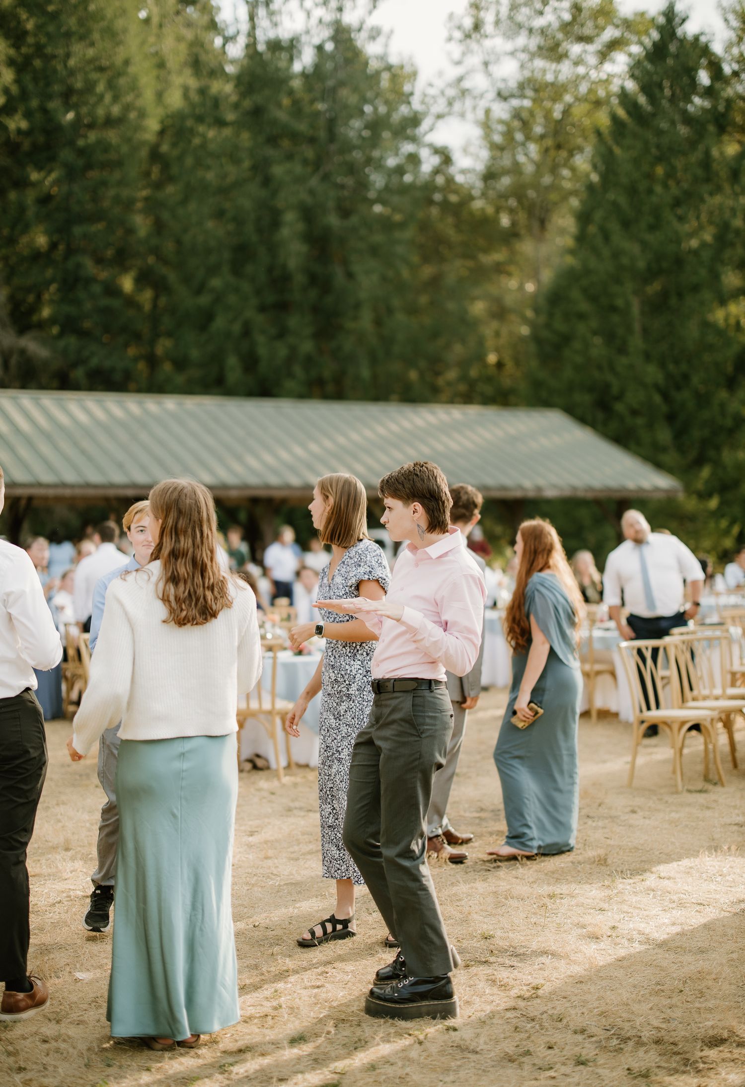 Guests dancing outdoors at a rustic wedding reception with tall trees and a covered pavilion in the background.