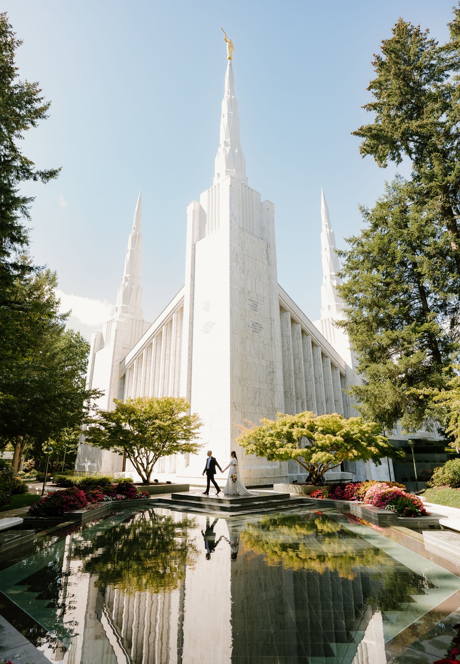 Couple holds hands on steps of white LDS temple with reflection pool and golden angel Moroni spire.