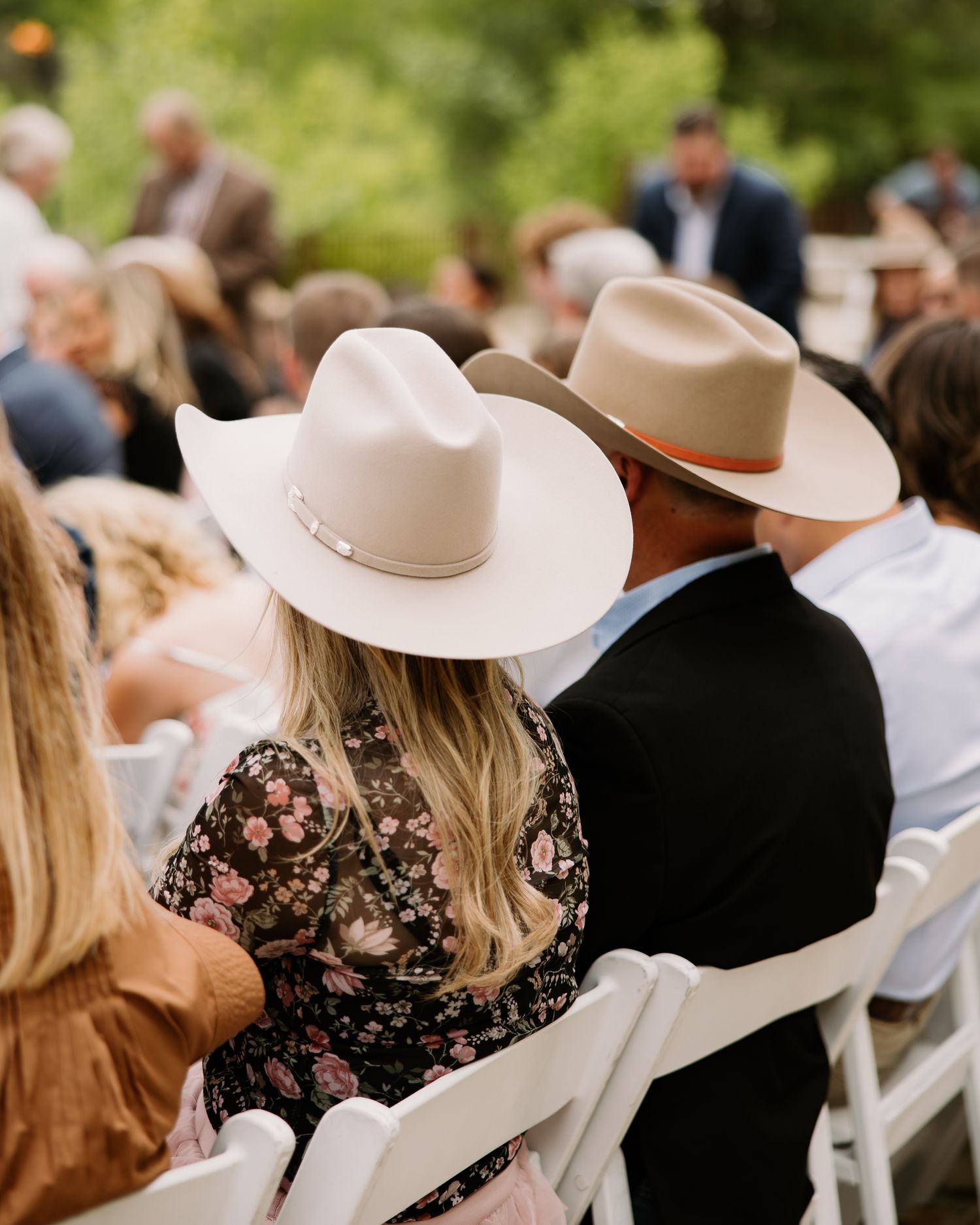 Couple wearing matching western cowboy hats seated at an outdoor wedding ceremony among other guests.