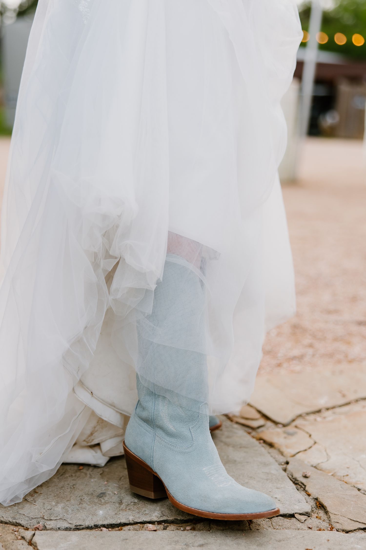 Bride wearing light blue suede cowboy boots beneath white wedding dress on stone outdoor venue floor.