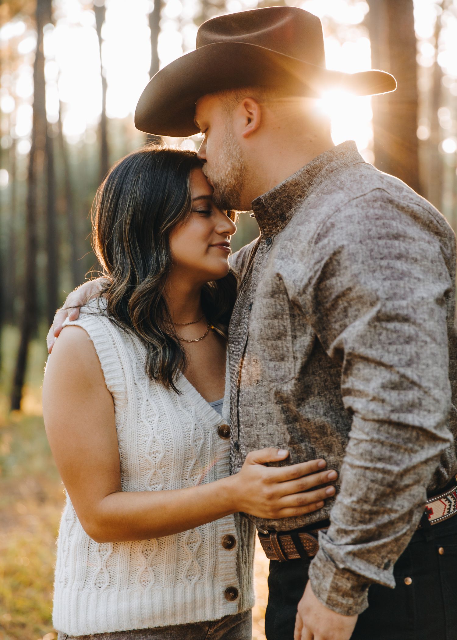 Couple sharing an intimate moment in a sunlit forest, man wearing a cowboy hat at golden hour.