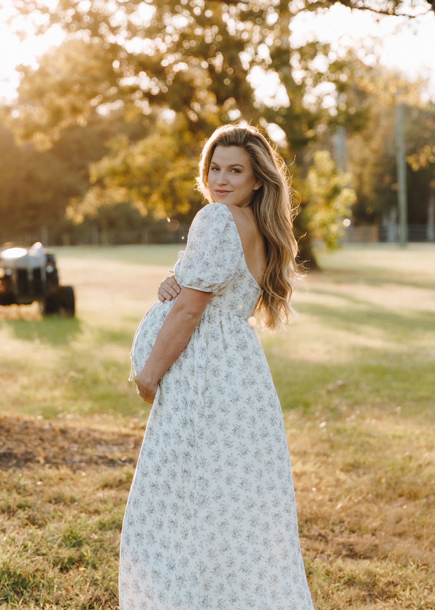 Pregnant woman in floral puff-sleeve dress cradles bump at golden hour in scenic outdoor setting.