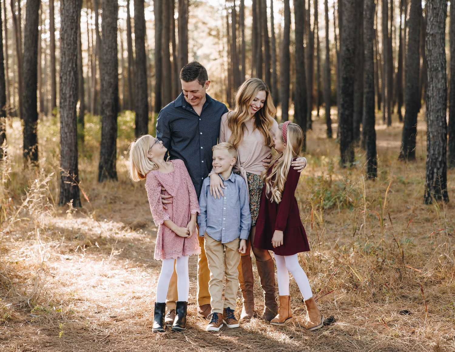Family of five smiling together on sunlit forest path among tall pine trees.