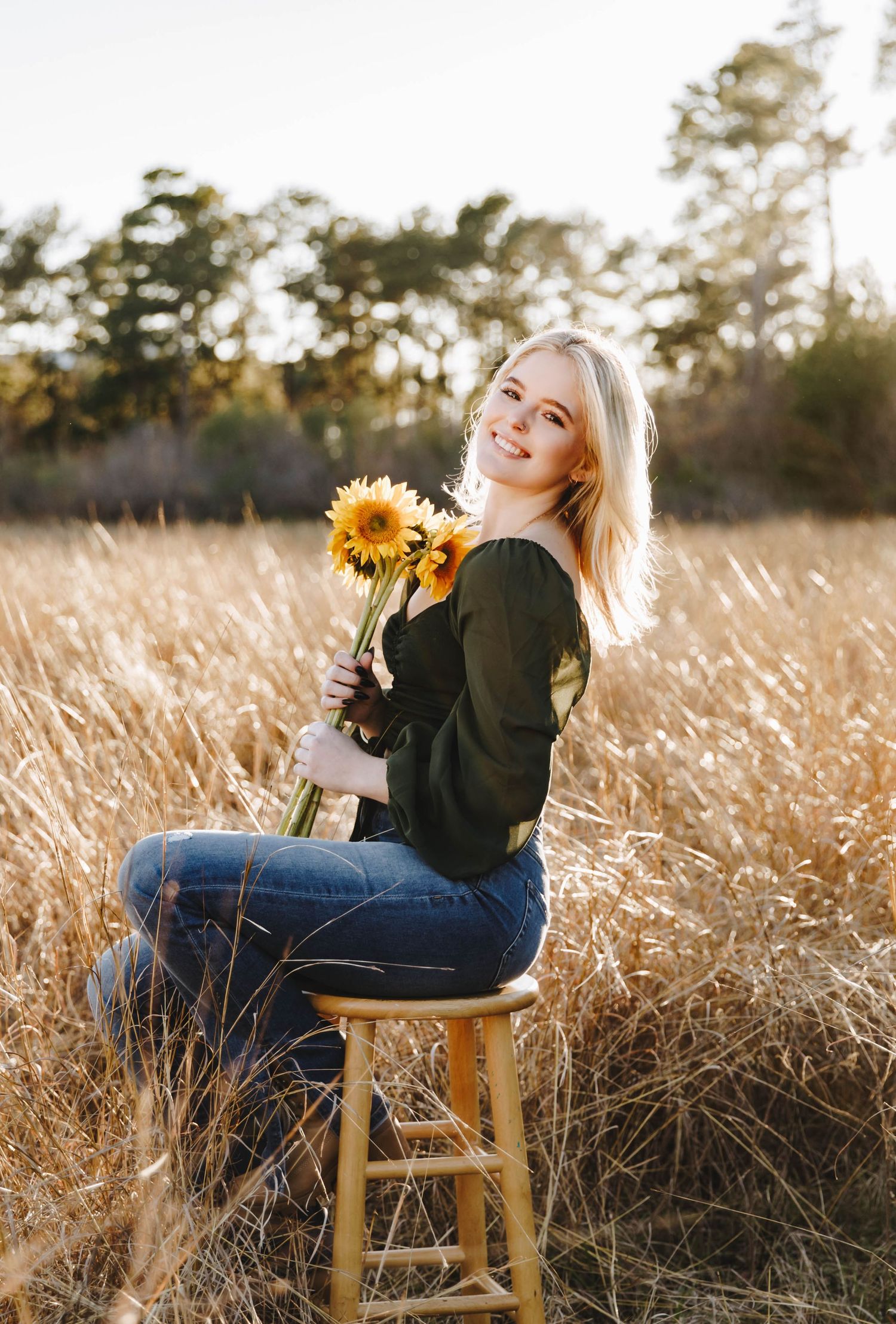 Smiling blonde woman sitting on a stool holding sunflowers in a golden field at sunset.