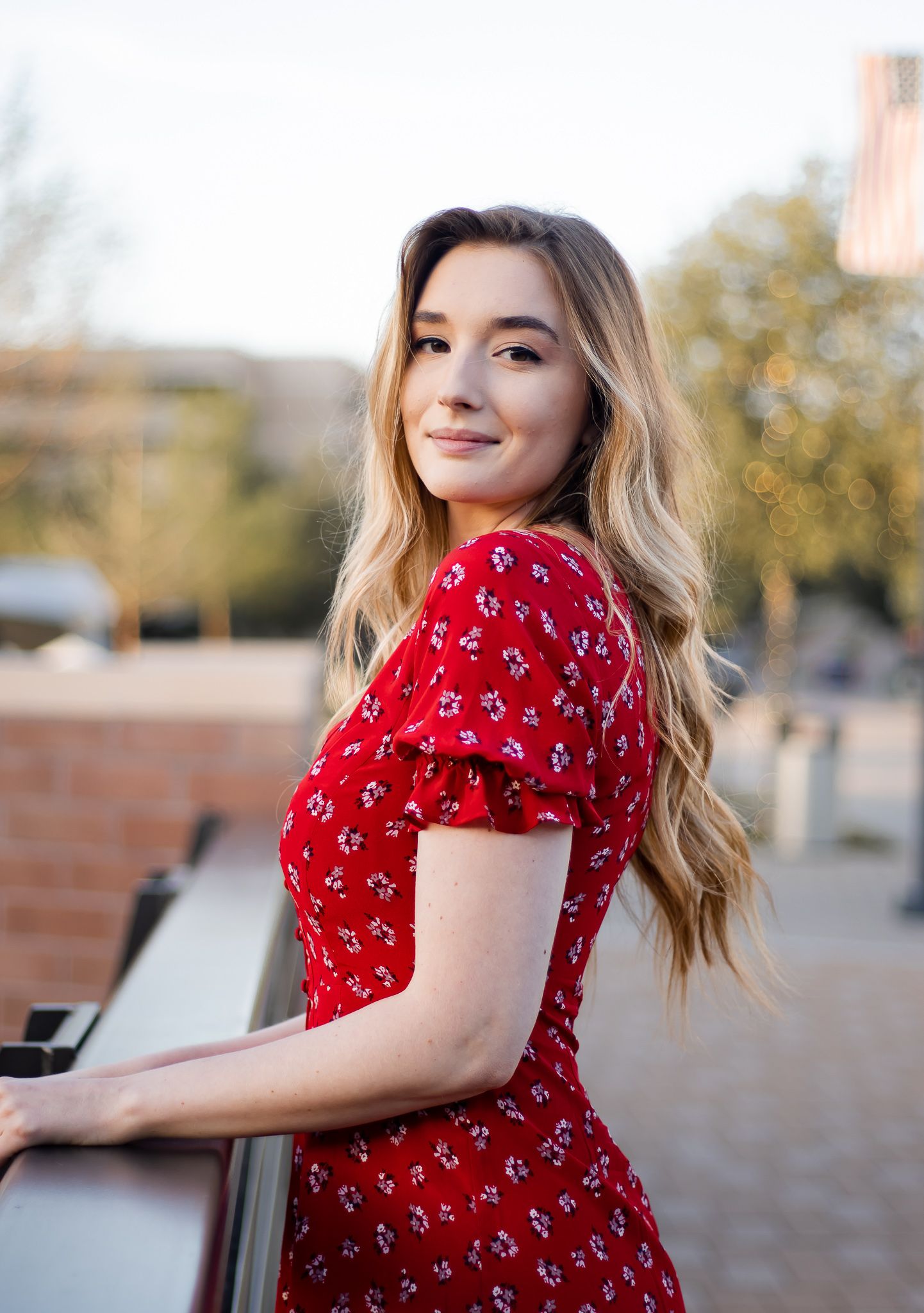 Young woman in red floral dress leans on railing with blurred urban background.