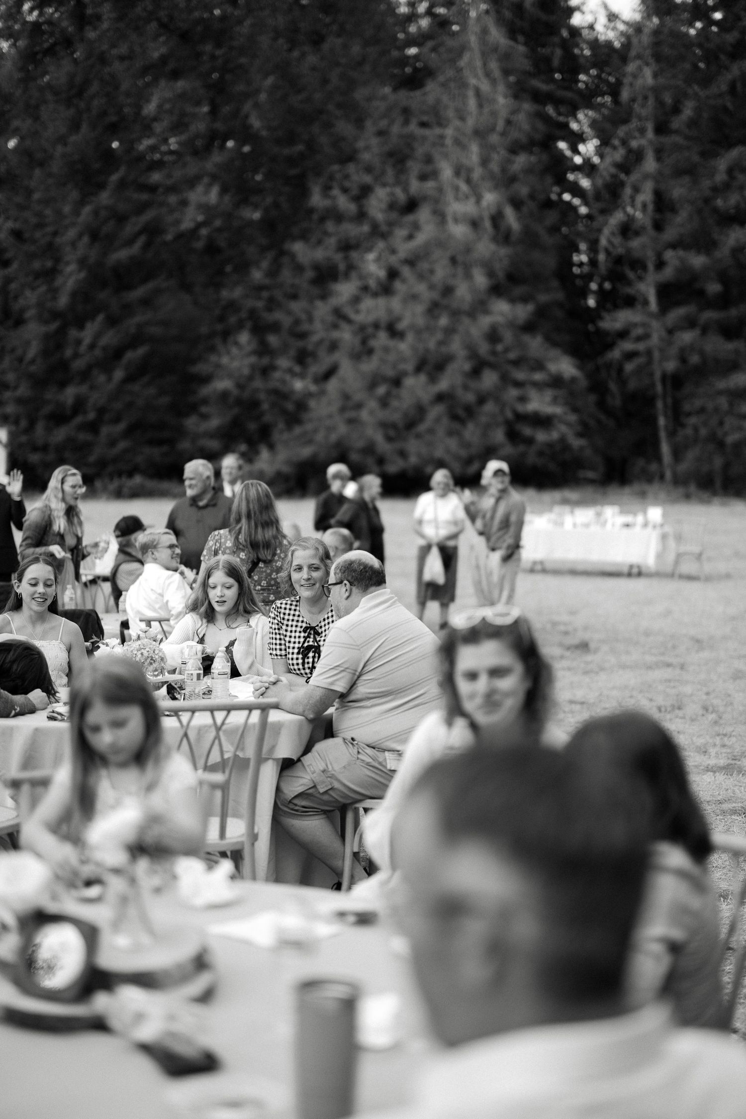 Black and white photo of wedding reception guests dining at long outdoor tables among tall trees.
