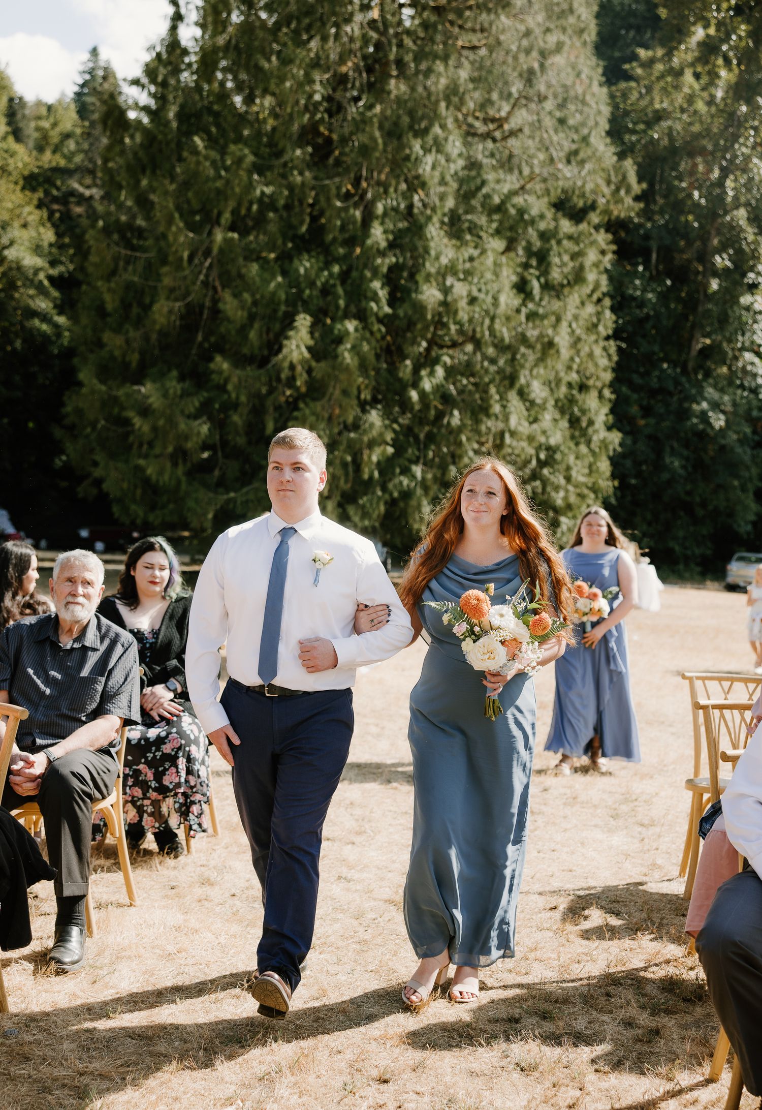 Bridesmaid and groomsman walk down outdoor wedding aisle surrounded by seated guests and tall evergreen trees.
