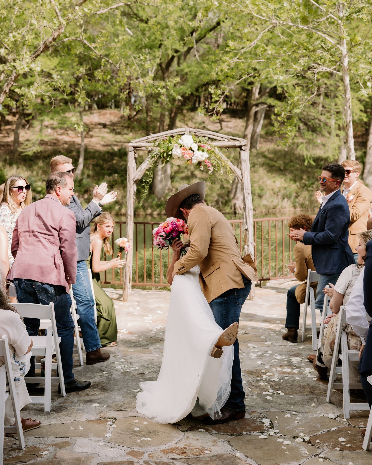Couple sharing first kiss at outdoor rustic wedding ceremony with floral arch, guests applauding on stone patio.