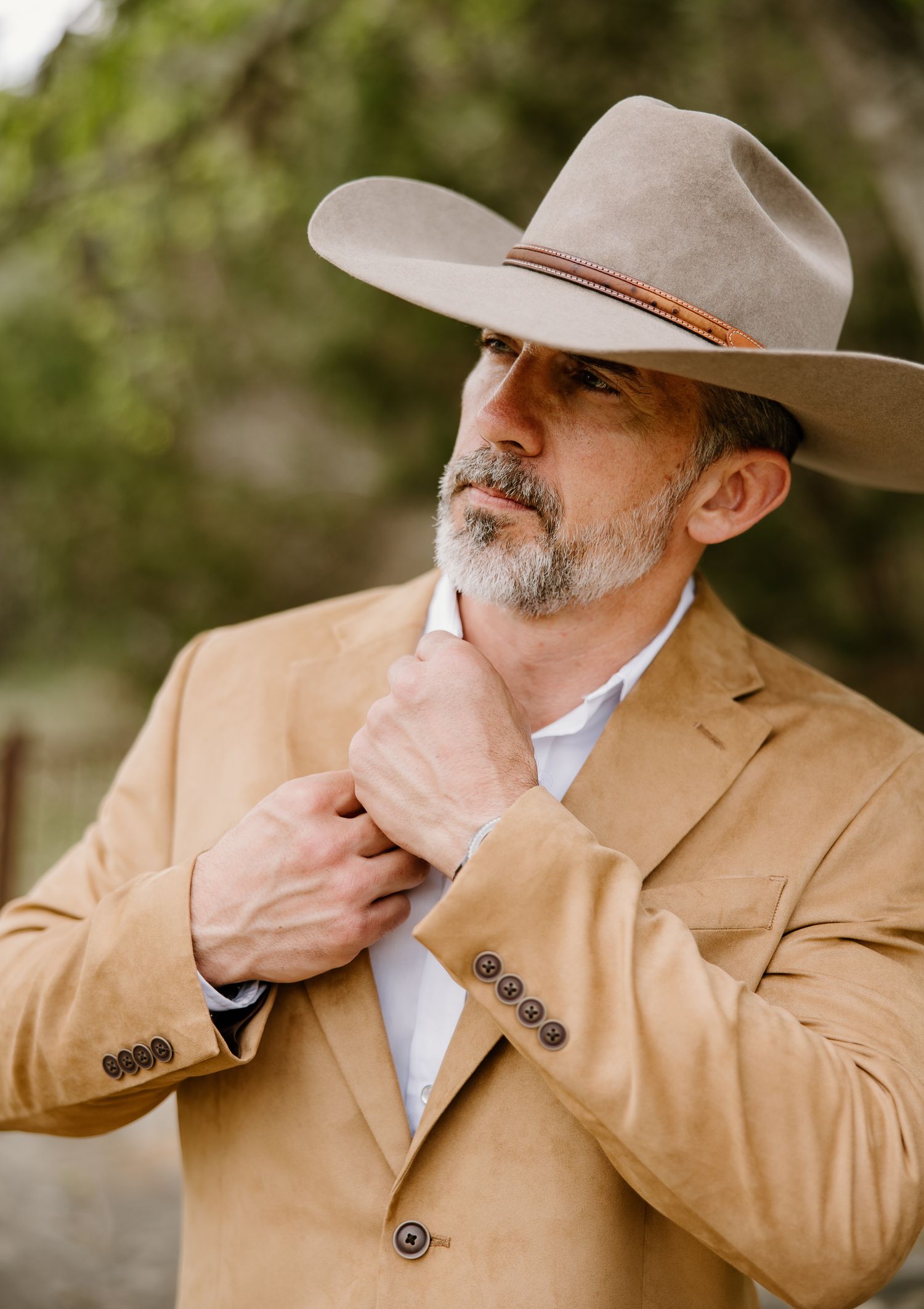 A middle-aged man with a gray beard wearing a tan cowboy hat and beige blazer adjusts his collar outdoors.
